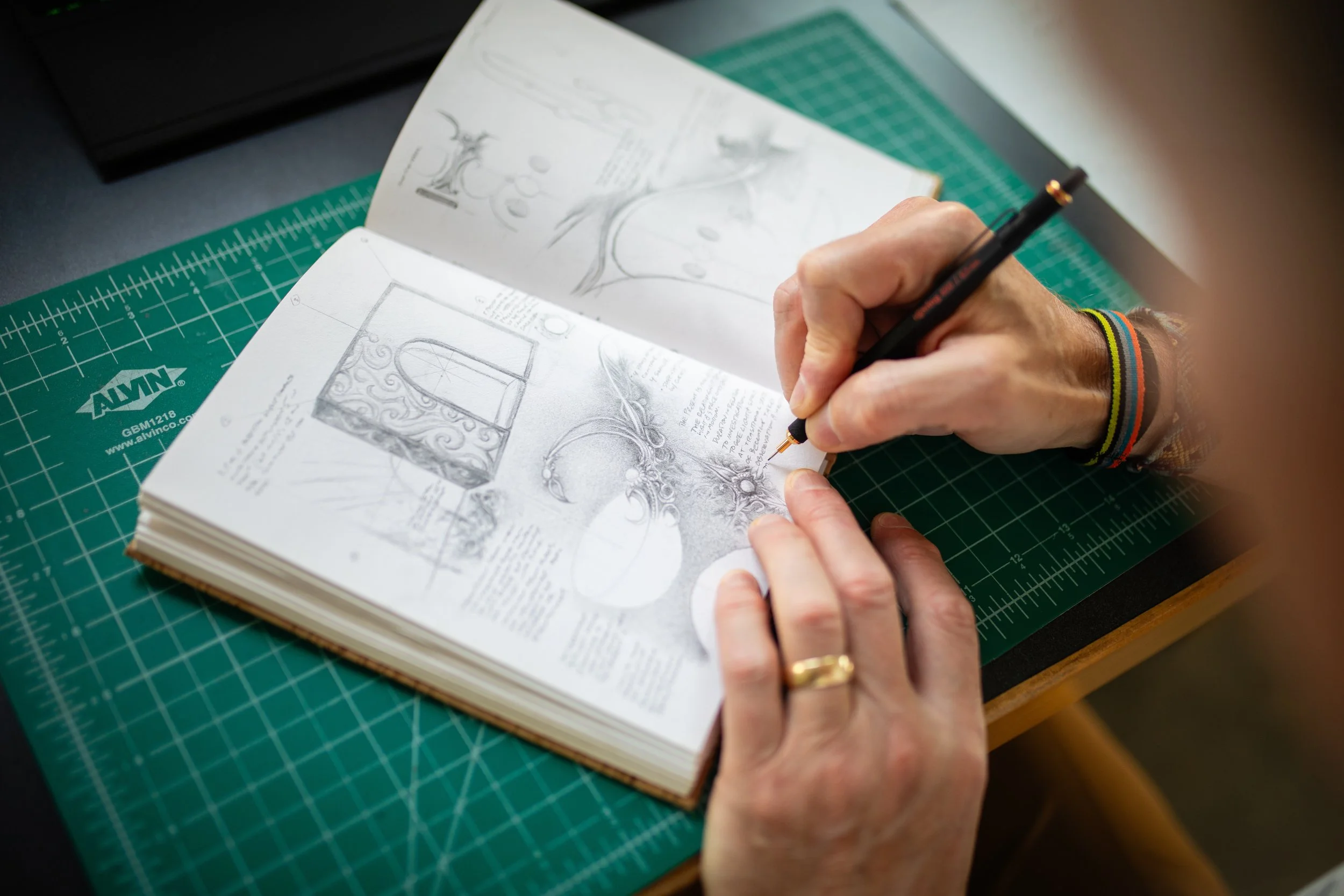 Person working on detailed jewelry sketches or design drawings at a worktable with art and design books open.