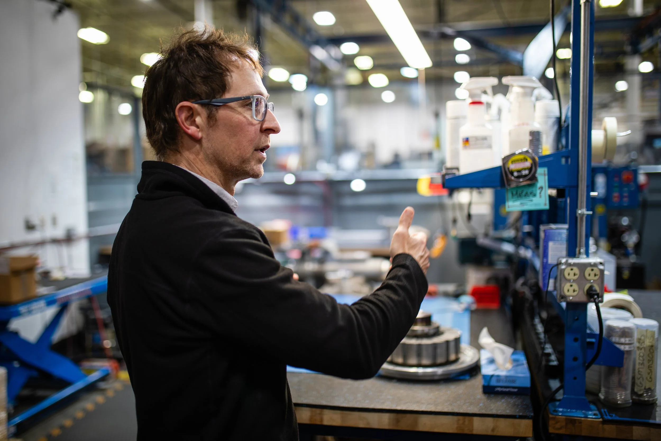 A man with glasses wearing a black jacket is in a workshop, pointing at a shelf with various bottles and small containers.