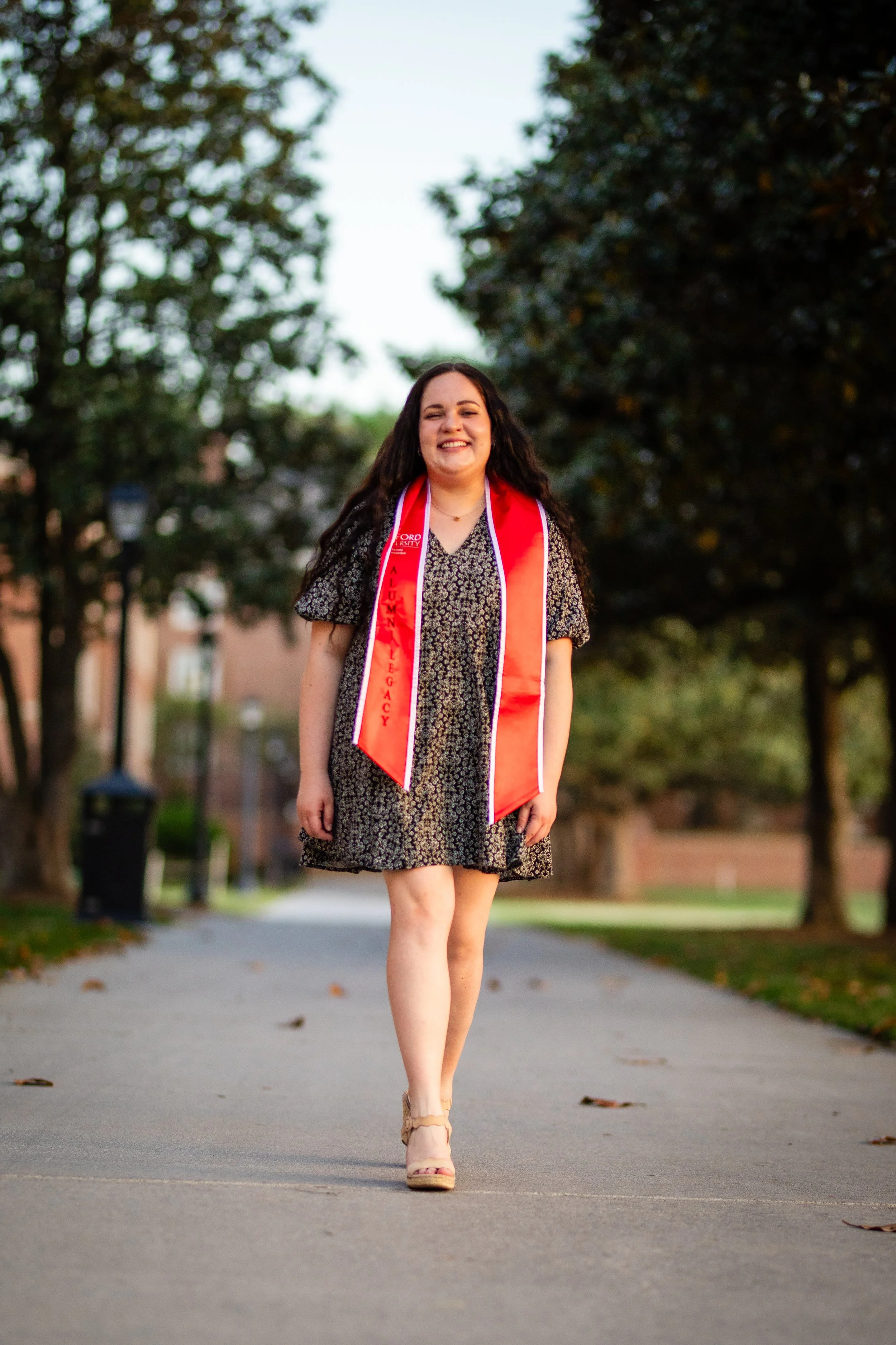 A young woman with long dark hair is walking outdoors on a paved path, wearing a black patterned dress, beige high-heeled sandals, and a red graduation stole that reads "Alumni Legacy." She is smiling and appears happy, with trees and a clear sky in 