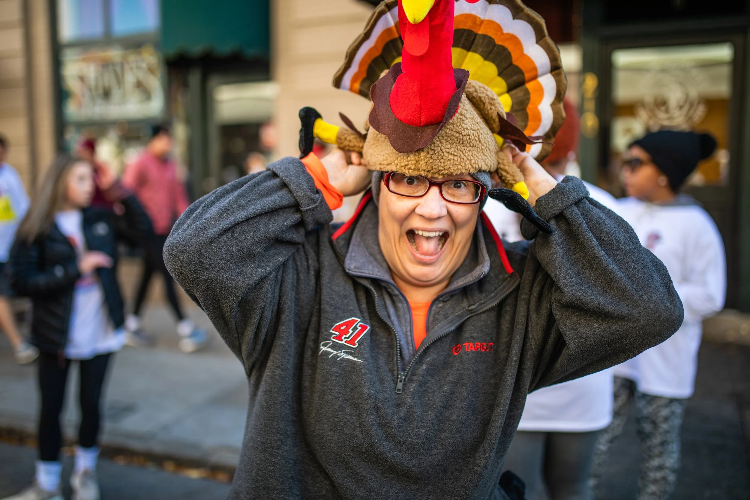 Person wearing a turkey hat and glasses, smiling and holding the turkey hat on their head, with a group of people in the background.