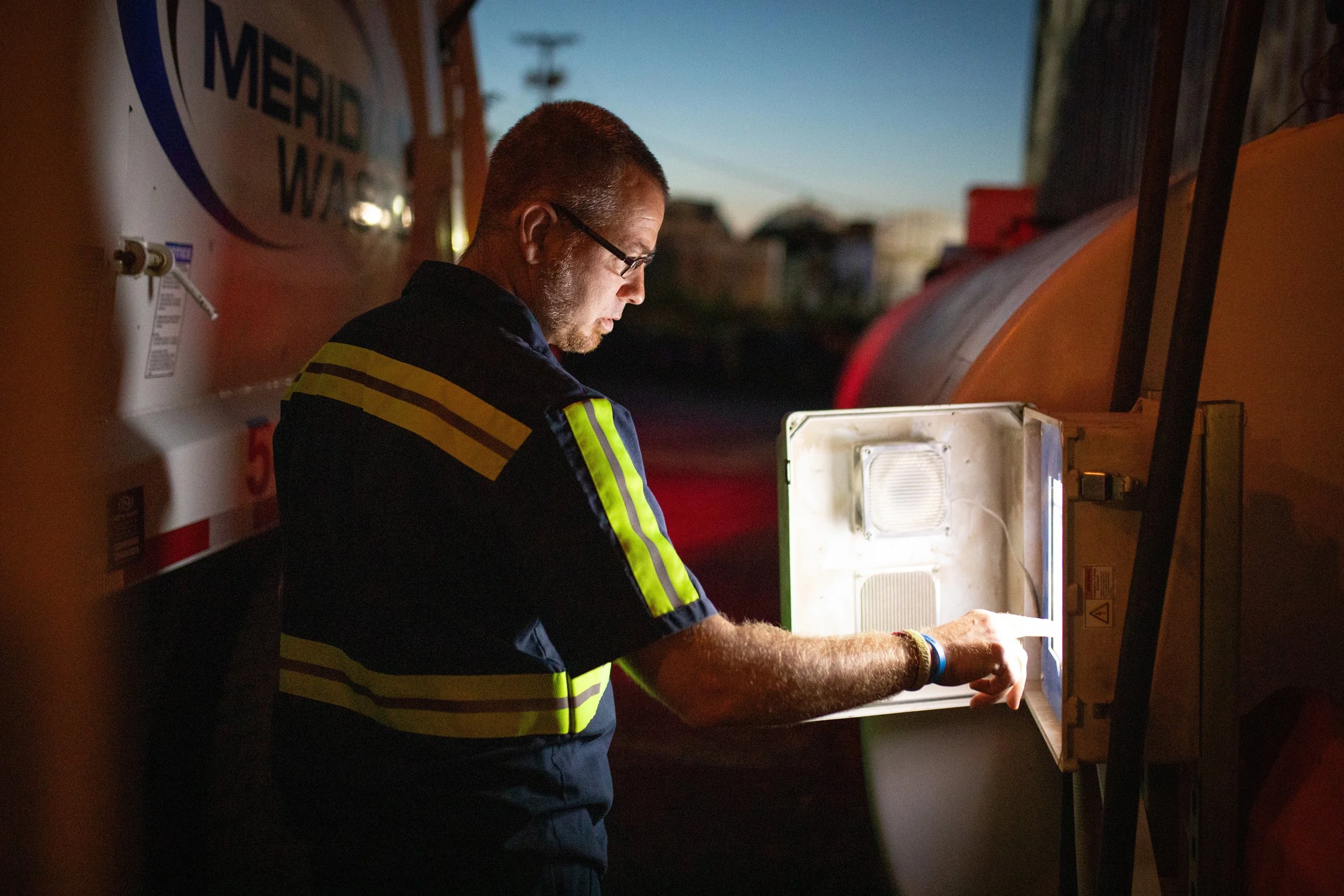 A man in a uniform inspecting a small refrigeration unit outside during dusk or early evening.