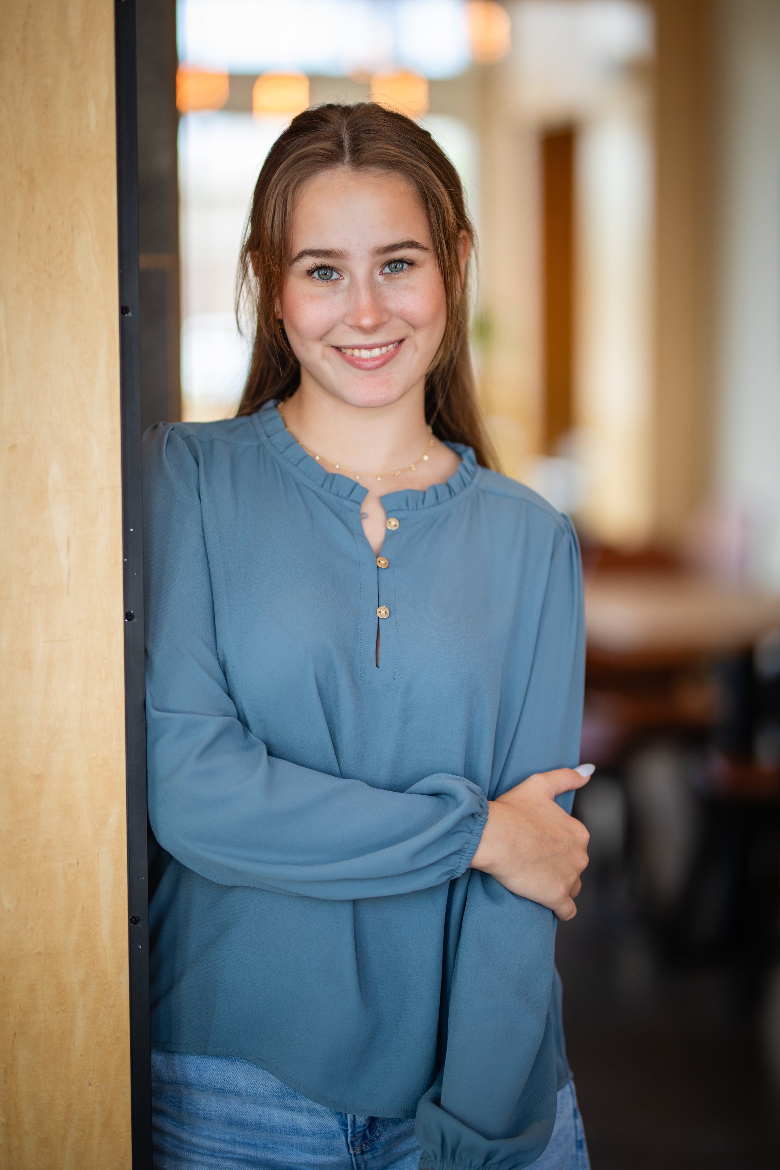 A young woman with long brown hair and blue eyes is smiling and standing indoors, leaning against a wood-paneled wall with a blurred background.