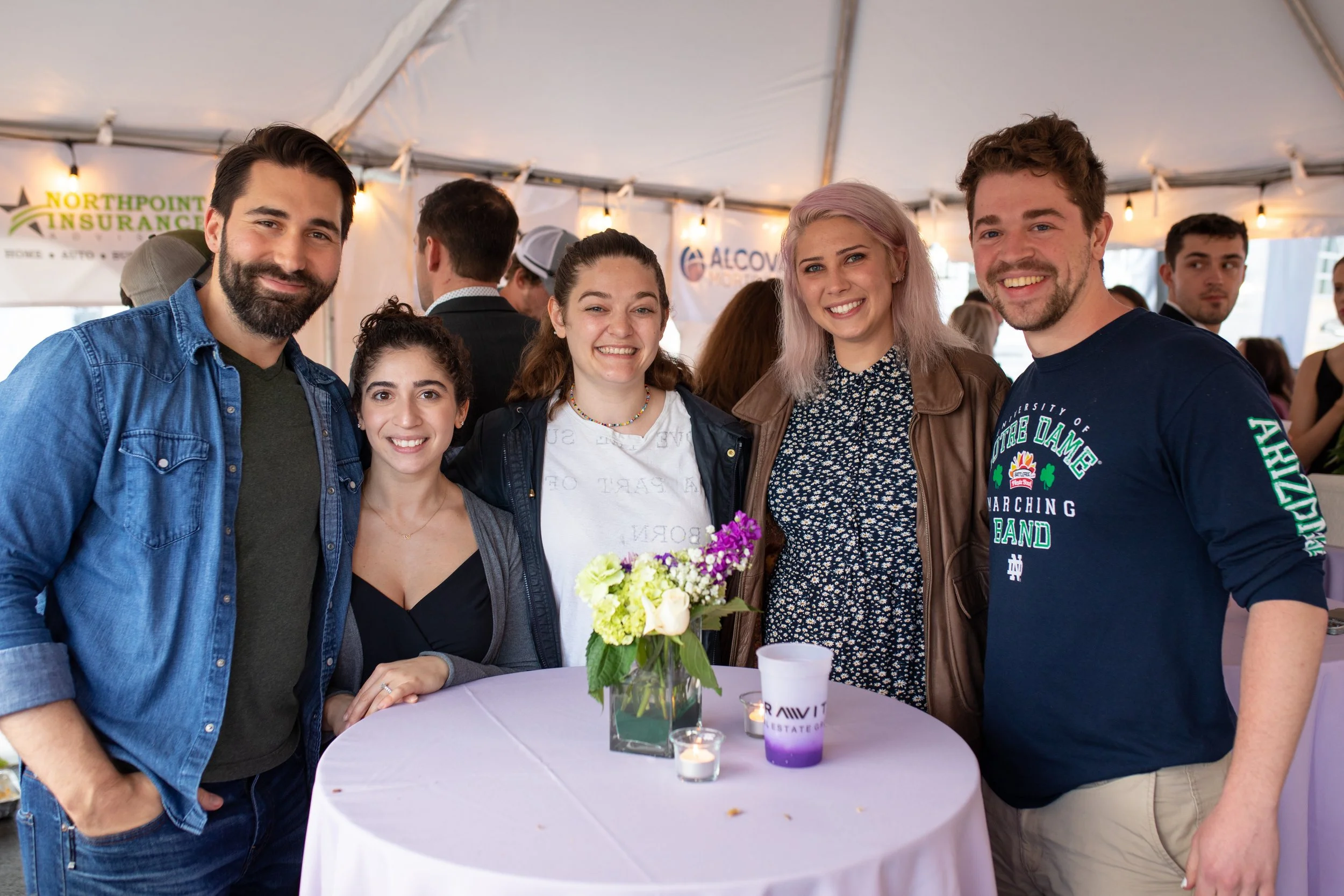 Group of five young people smiling at a social event, standing around a table with a flower arrangement and small candles under a white event tent.