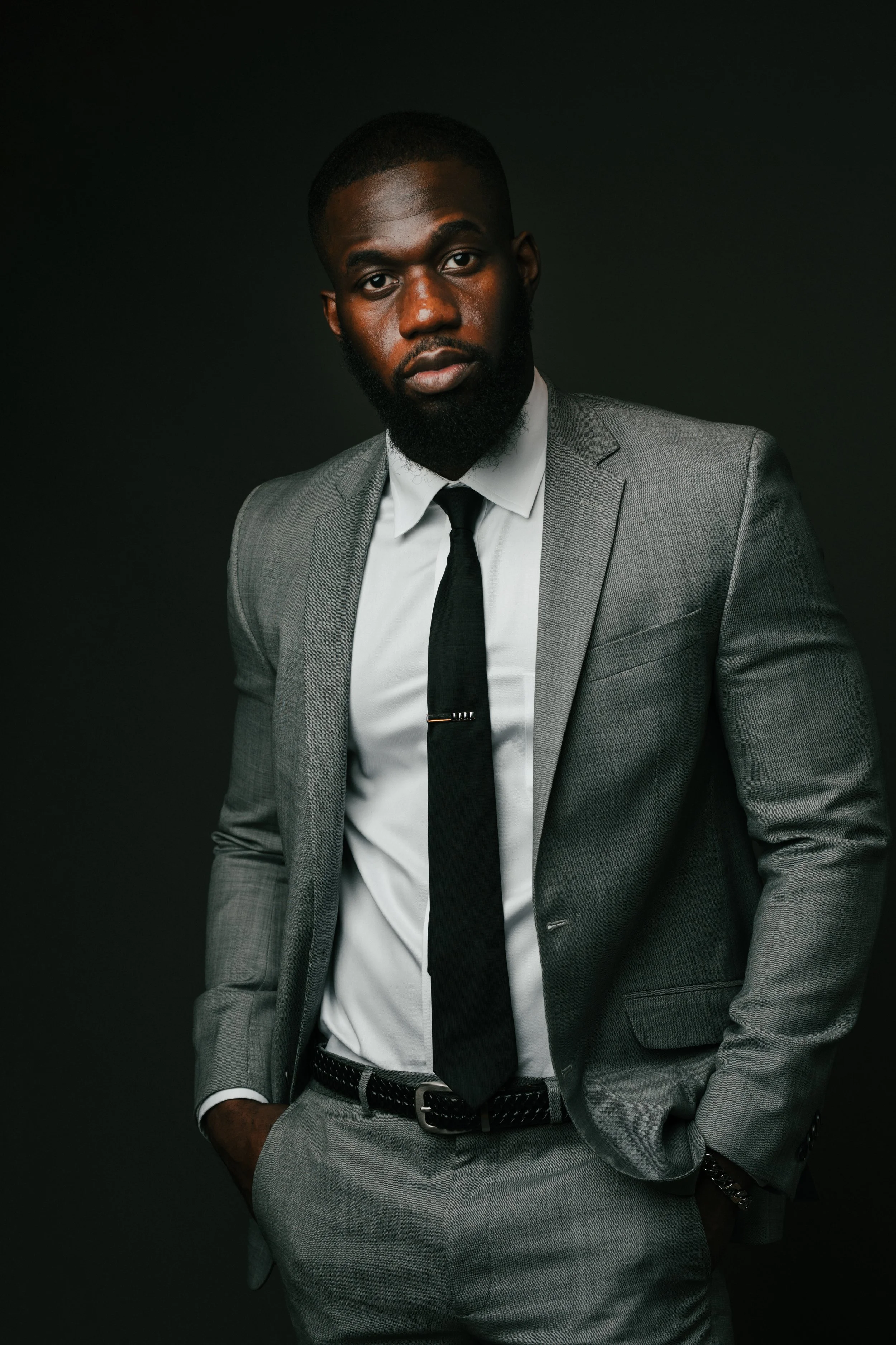 Young man with short hair and beard wearing a gray suit, white shirt, and black tie against a dark background.
