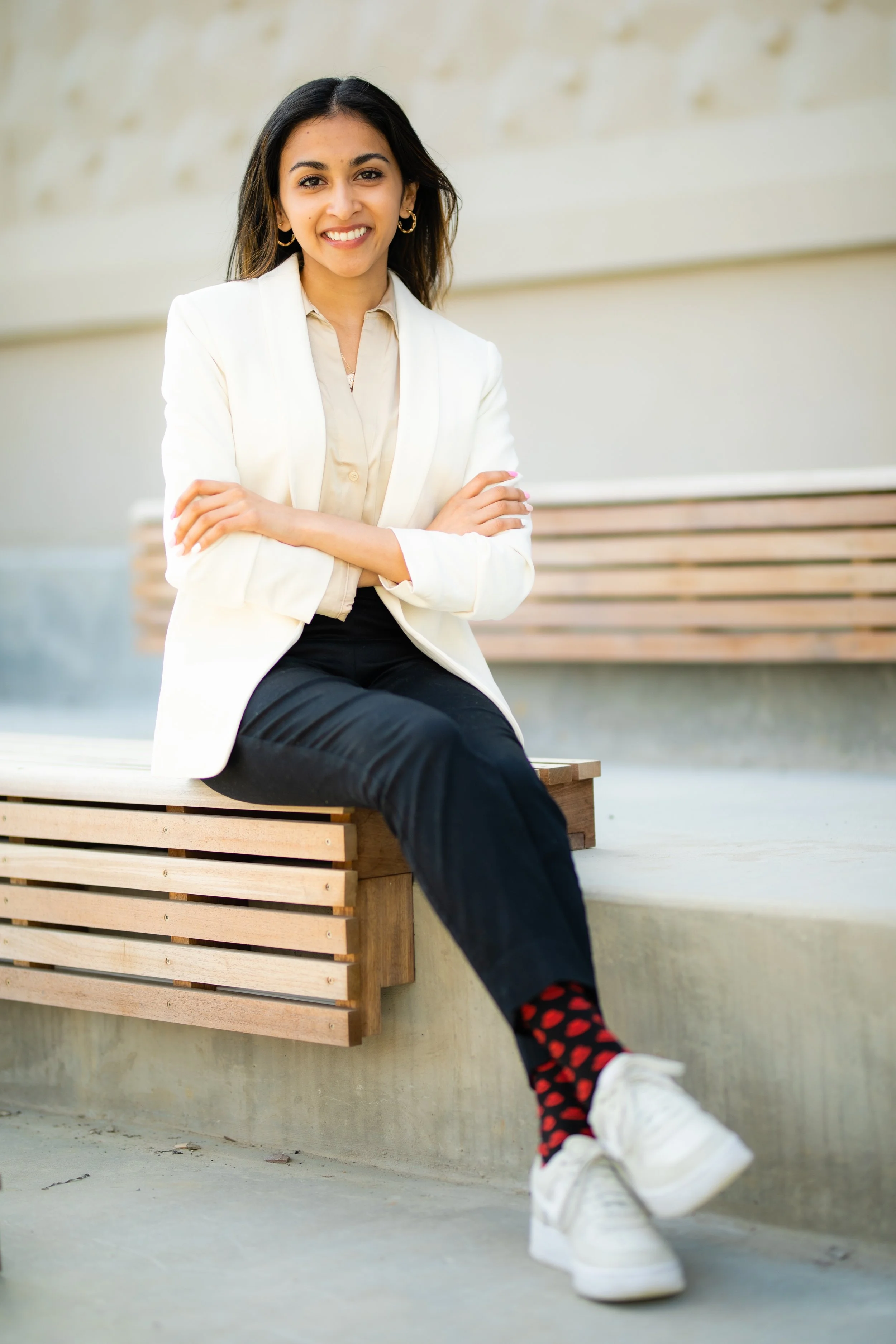 A woman sitting on a wooden bench outdoors, smiling with arms crossed, wearing a white blazer, beige blouse, black pants, colorful socks with heart patterns, and white sneakers.