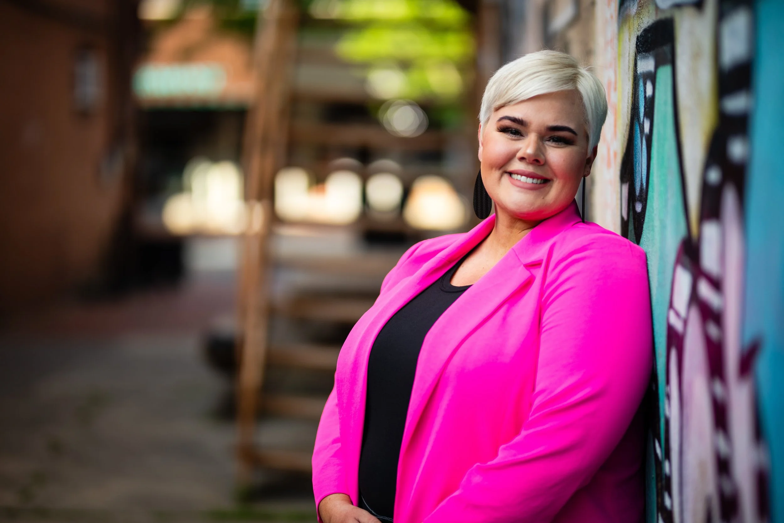 A woman with short platinum blonde hair wearing a bright pink blazer and black top, smiling and leaning against a colorful graffiti wall in an outdoor urban setting.