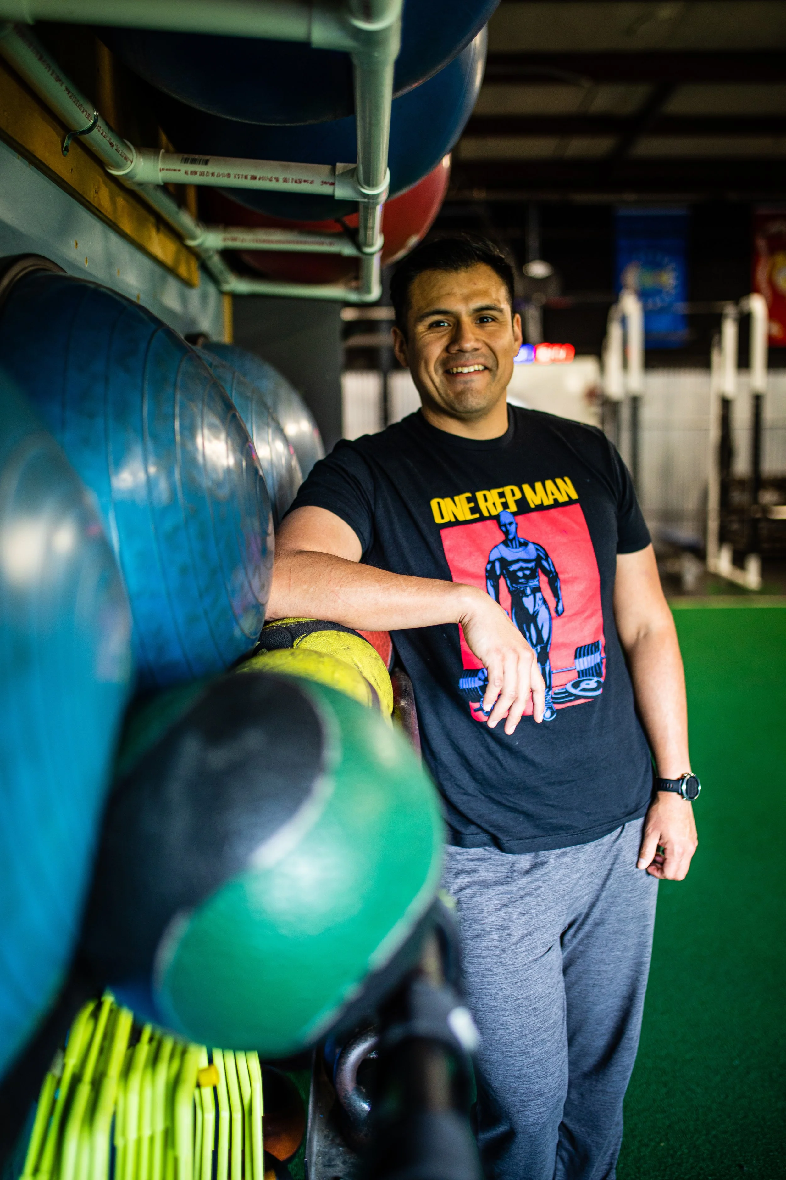A man in a black "One Rep Man" T-shirt, smiling and standing next to colorful medicine balls and rubber weights in a gym.