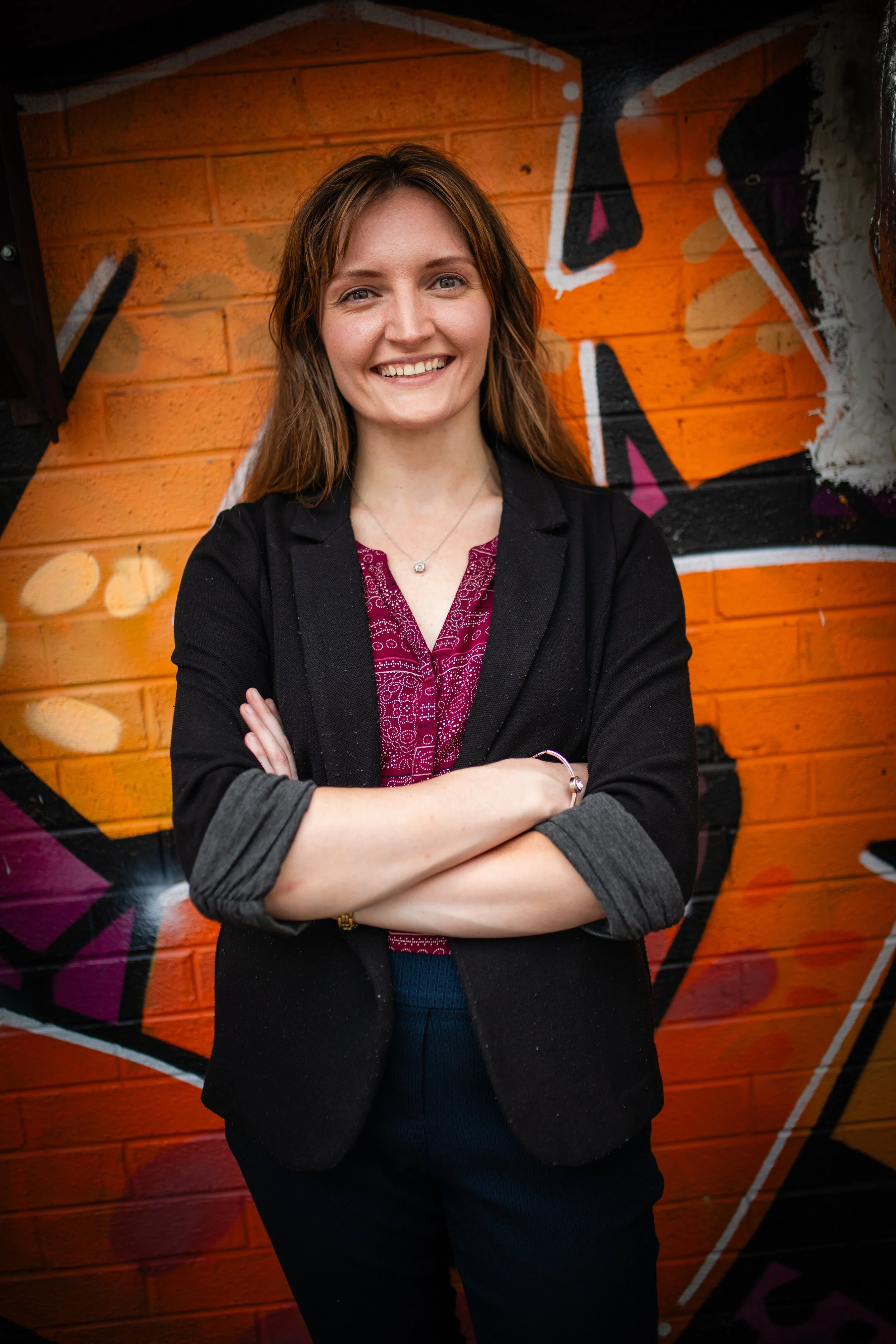 A woman with long brown hair, wearing a black blazer over a magenta patterned blouse, standing with her arms crossed in front of a vibrant graffiti mural on a brick wall.