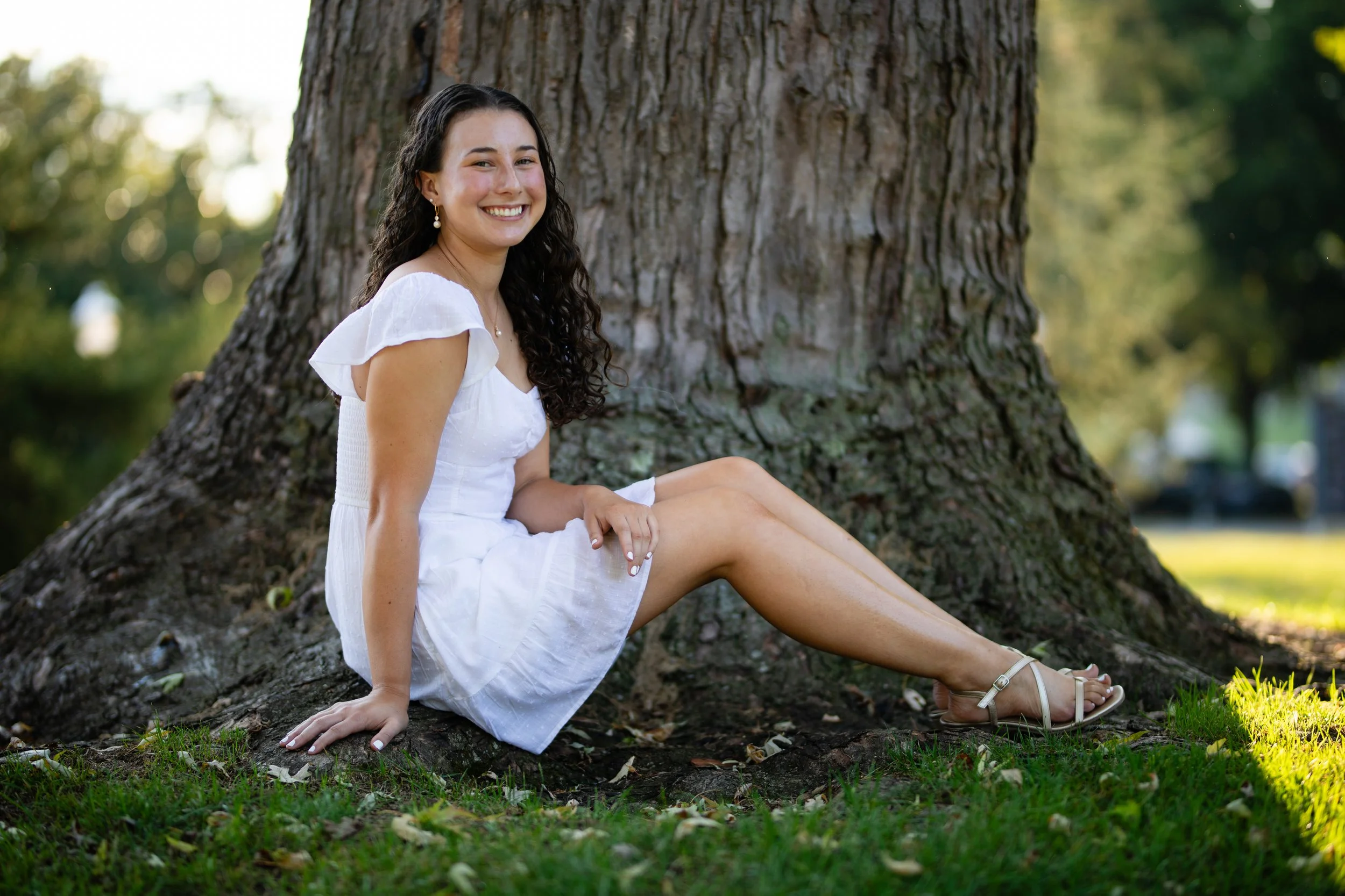 A young woman with long dark curly hair wearing a white dress and white sandals sitting on the grass and leaning against a large tree in a park, smiling at the camera.