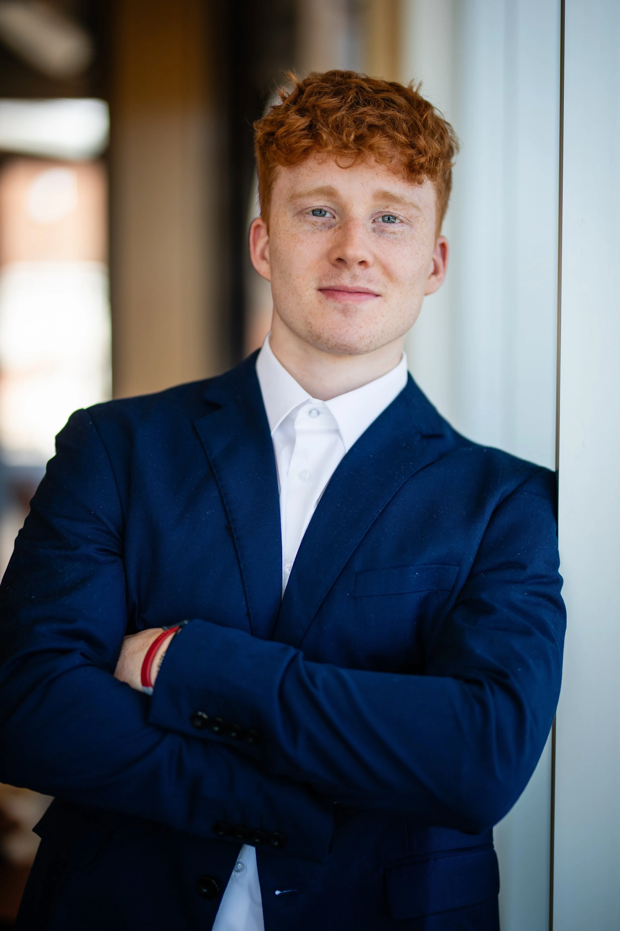Young man with red hair and freckles wearing a navy blue suit and white shirt, standing with arms crossed in front of a window.