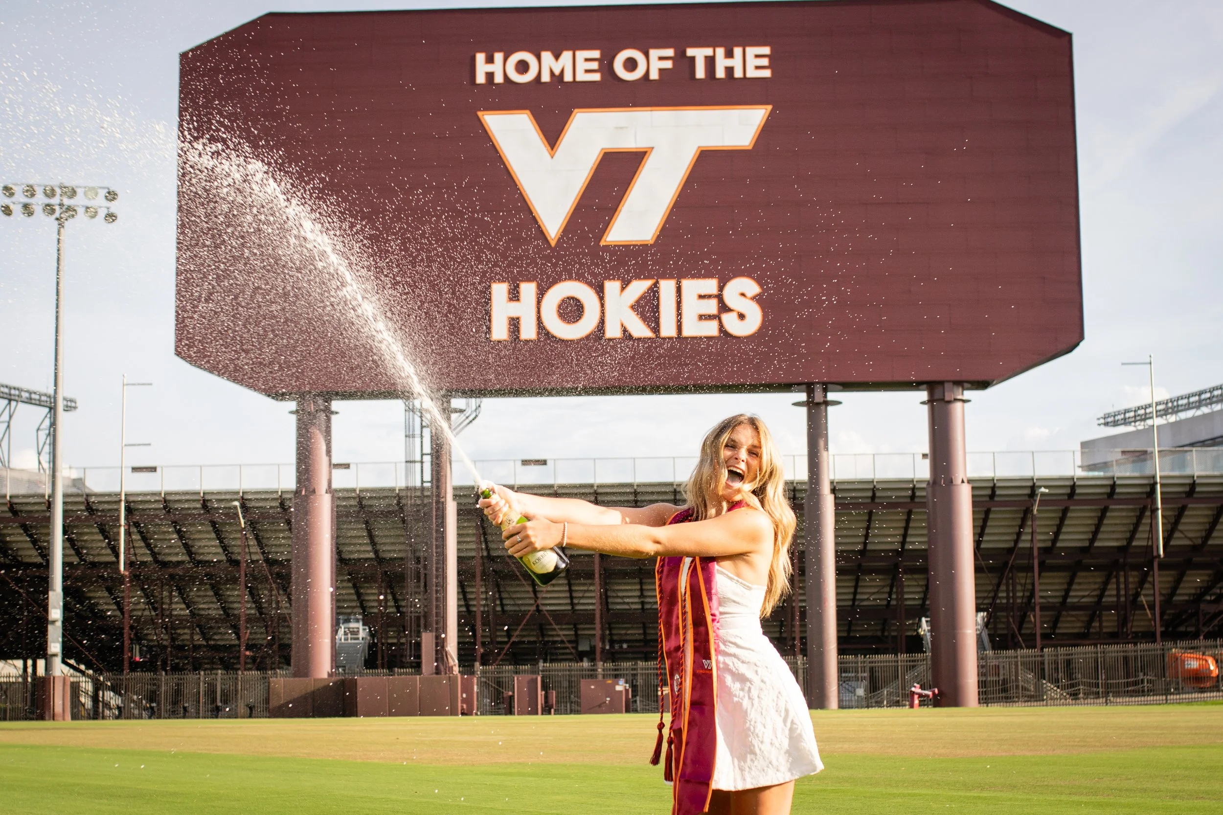 A woman celebrates on a sports field with a large scoreboard in the background that reads 'Home of the VT Hokies' and the Virginia Tech logo, while spraying champagne.