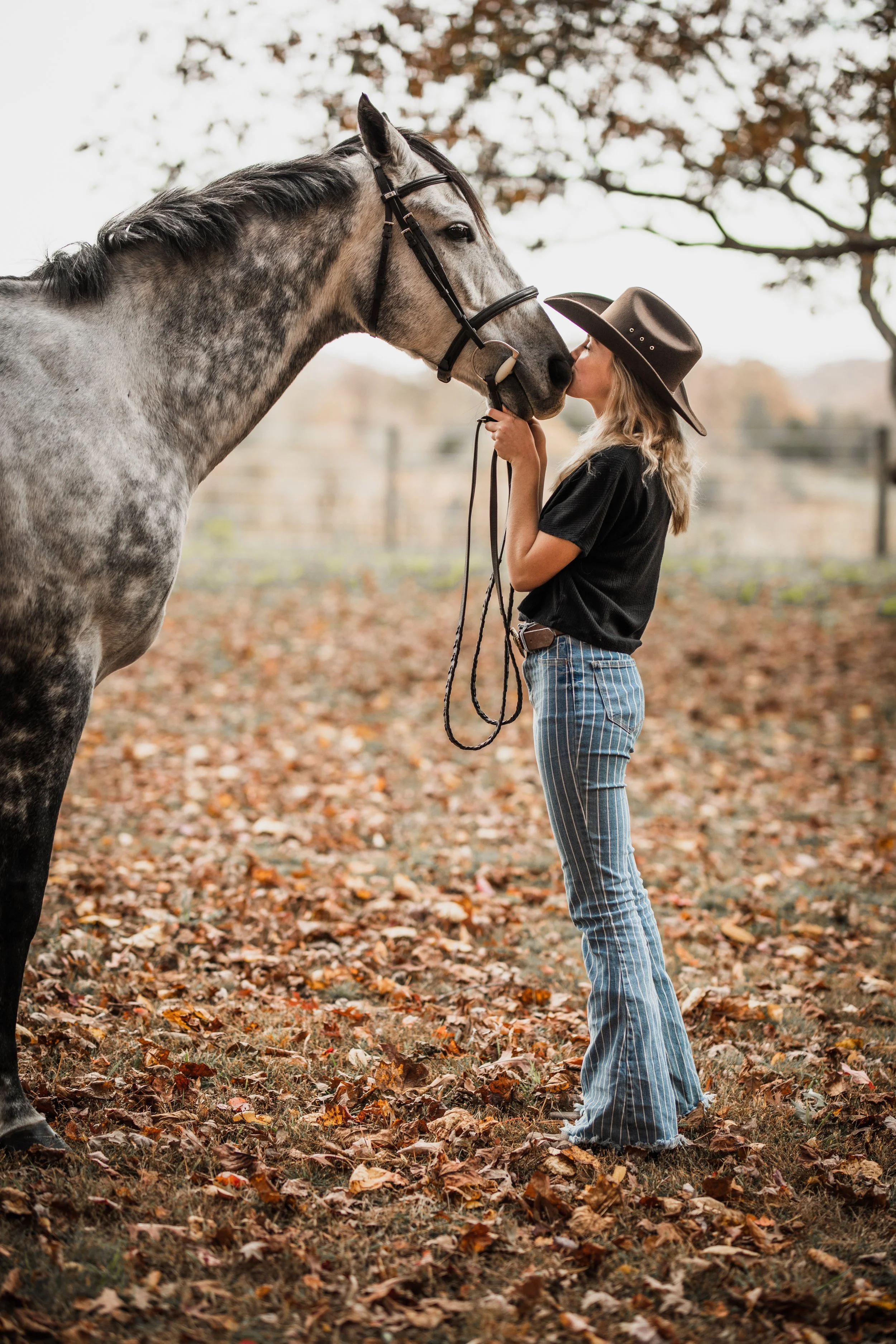 A woman wearing a cowboy hat and striped blue jeans kisses a gray horse on the nose in a fall outdoor setting.