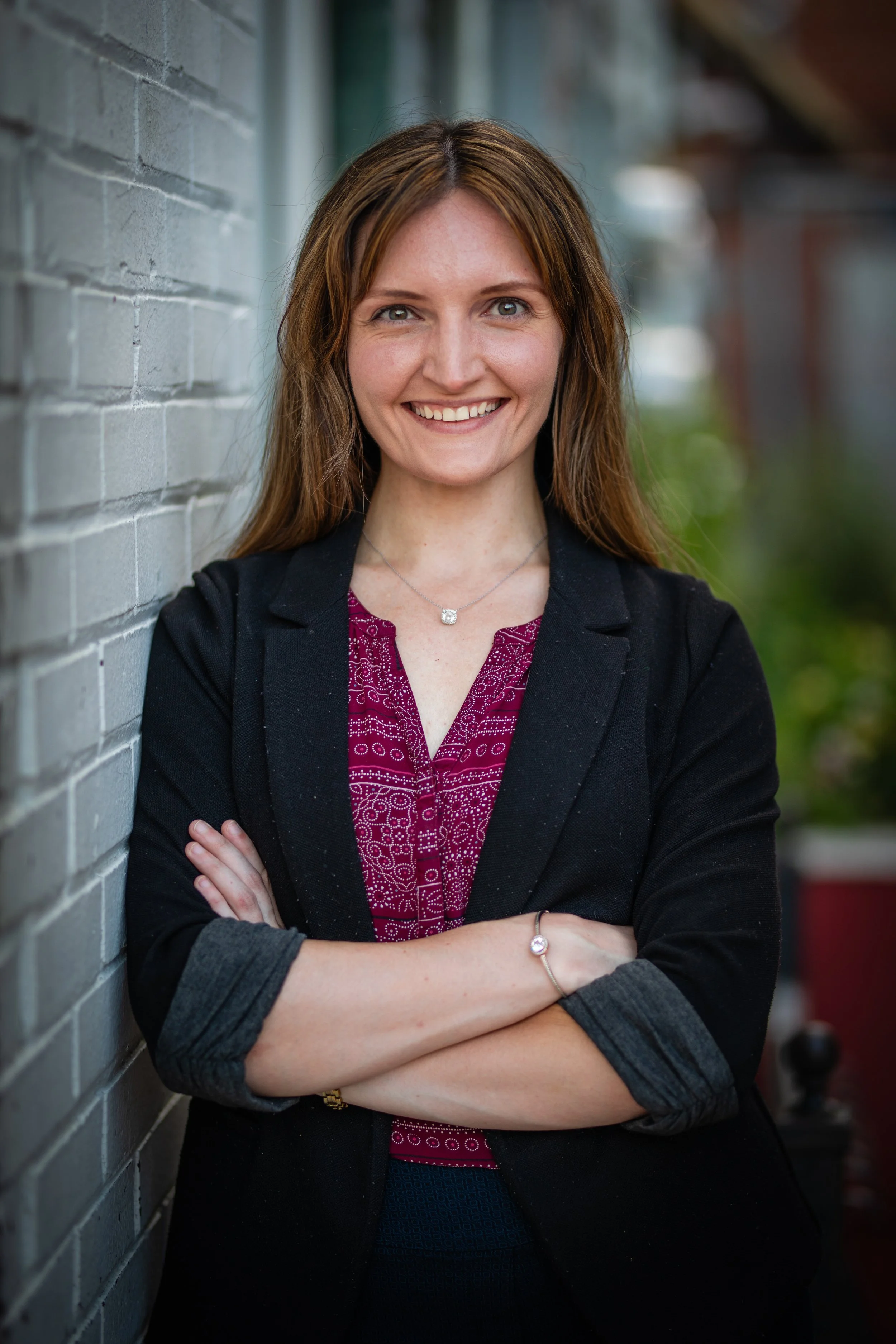 A woman with long brown hair smiling, standing with arms crossed against a brick wall outdoors.