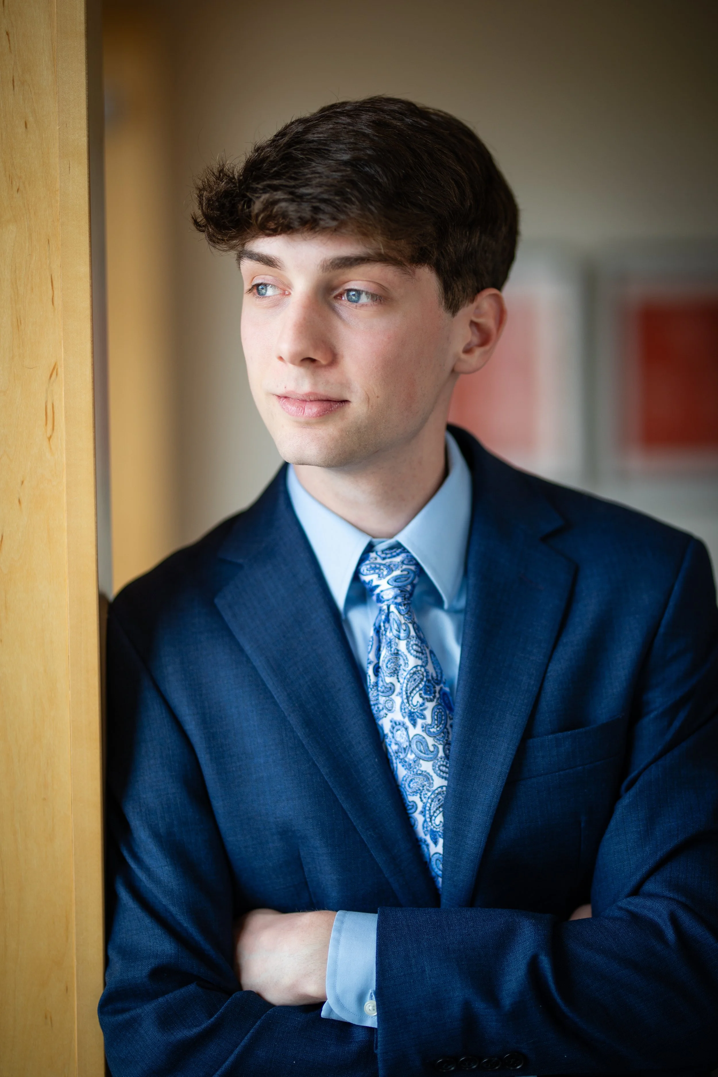 Young man in a navy suit and blue tie standing with arms crossed near a wooden wall, gazing thoughtfully out of the frame.