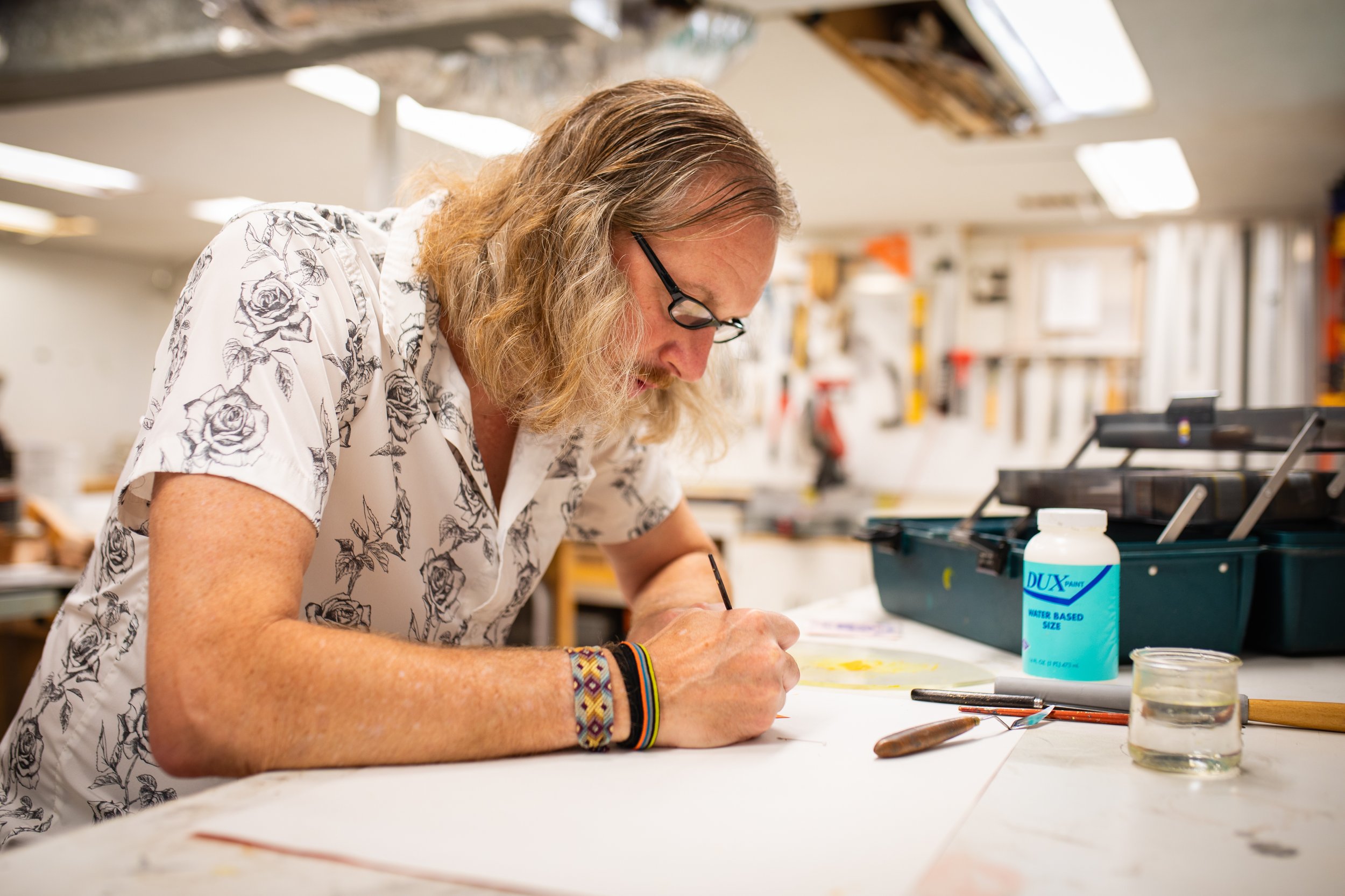 A person with long blonde hair wearing glasses and a white shirt with black roses is working on an art project in a workshop. They are leaning over a table, drawing or painting on paper.