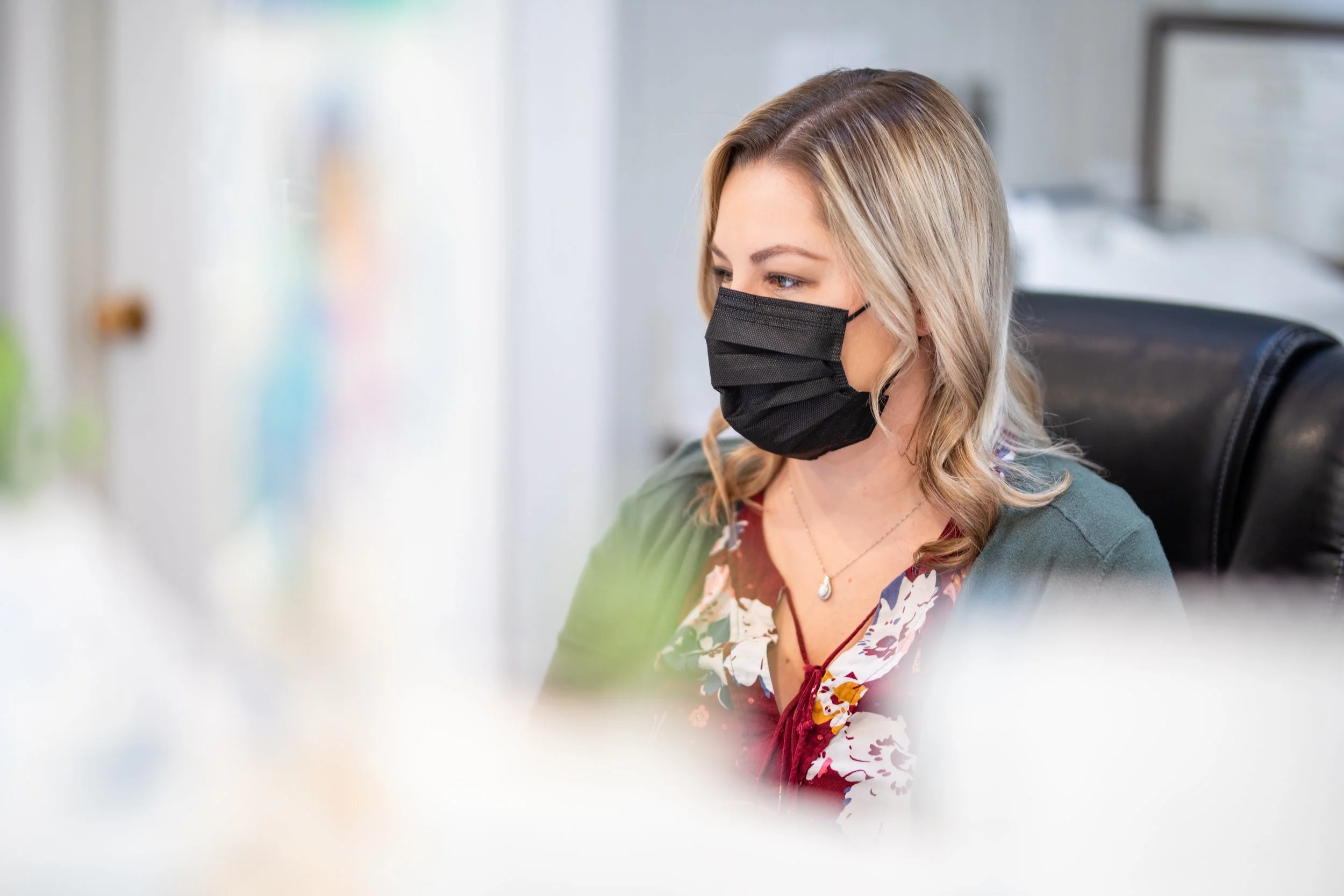 A woman with blonde hair wearing a black face mask, sitting in a modern office chair, working at a desk with a blurred background.