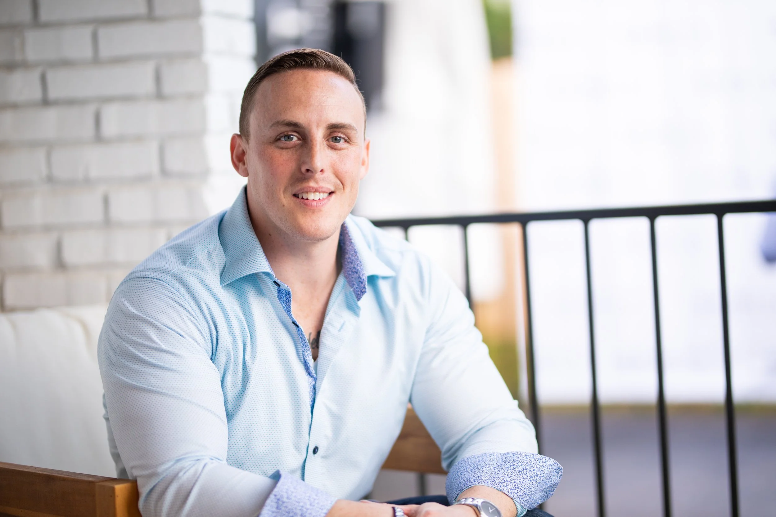 A man with short light-brown hair and light skin sitting at a table, smiling at the camera, wearing a light blue dress shirt with a patterned collar, inside a room with a white brick wall and a black railing in the background.