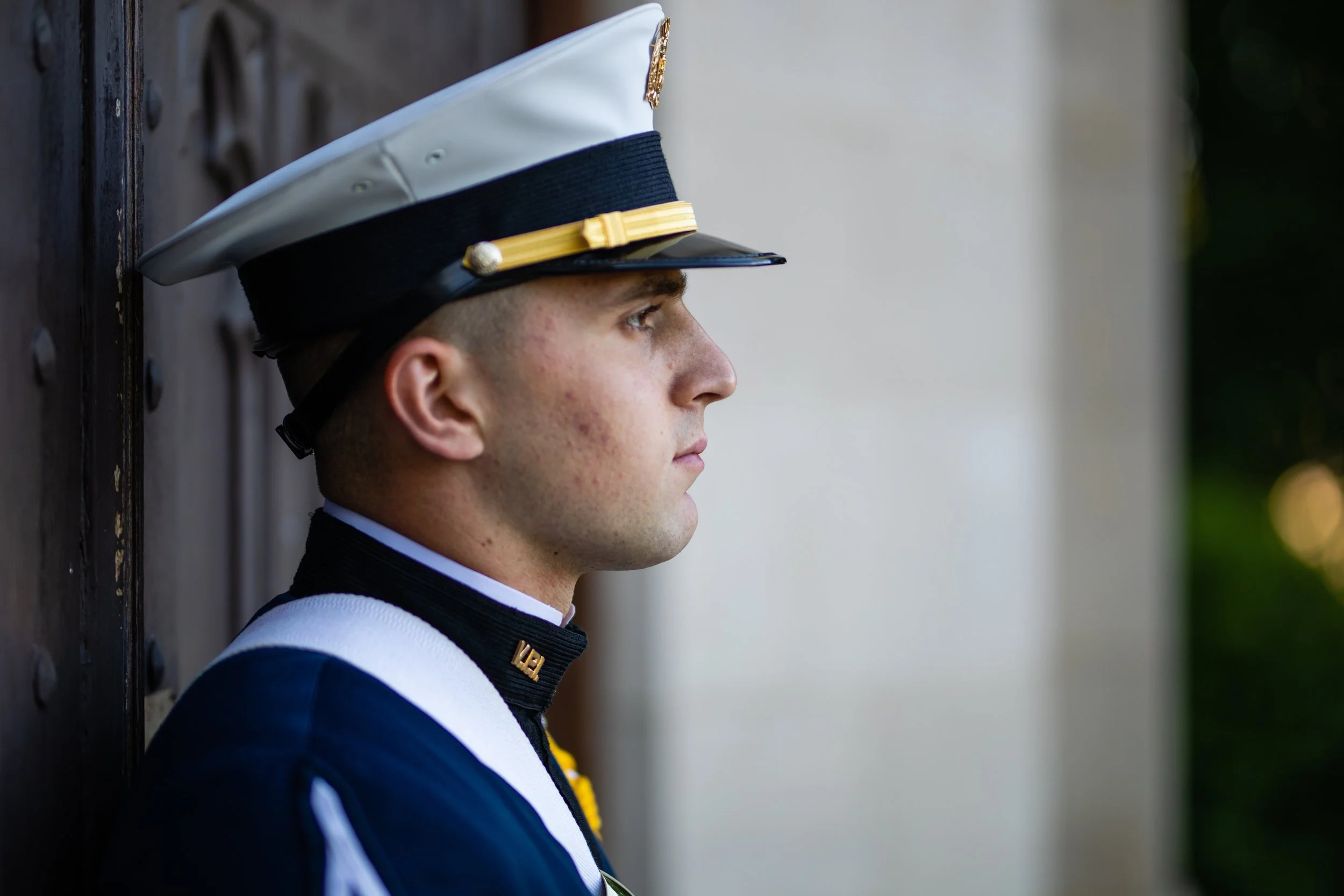 A young man in a United States Marine Corps dress uniform, standing in profile against a wooden background.