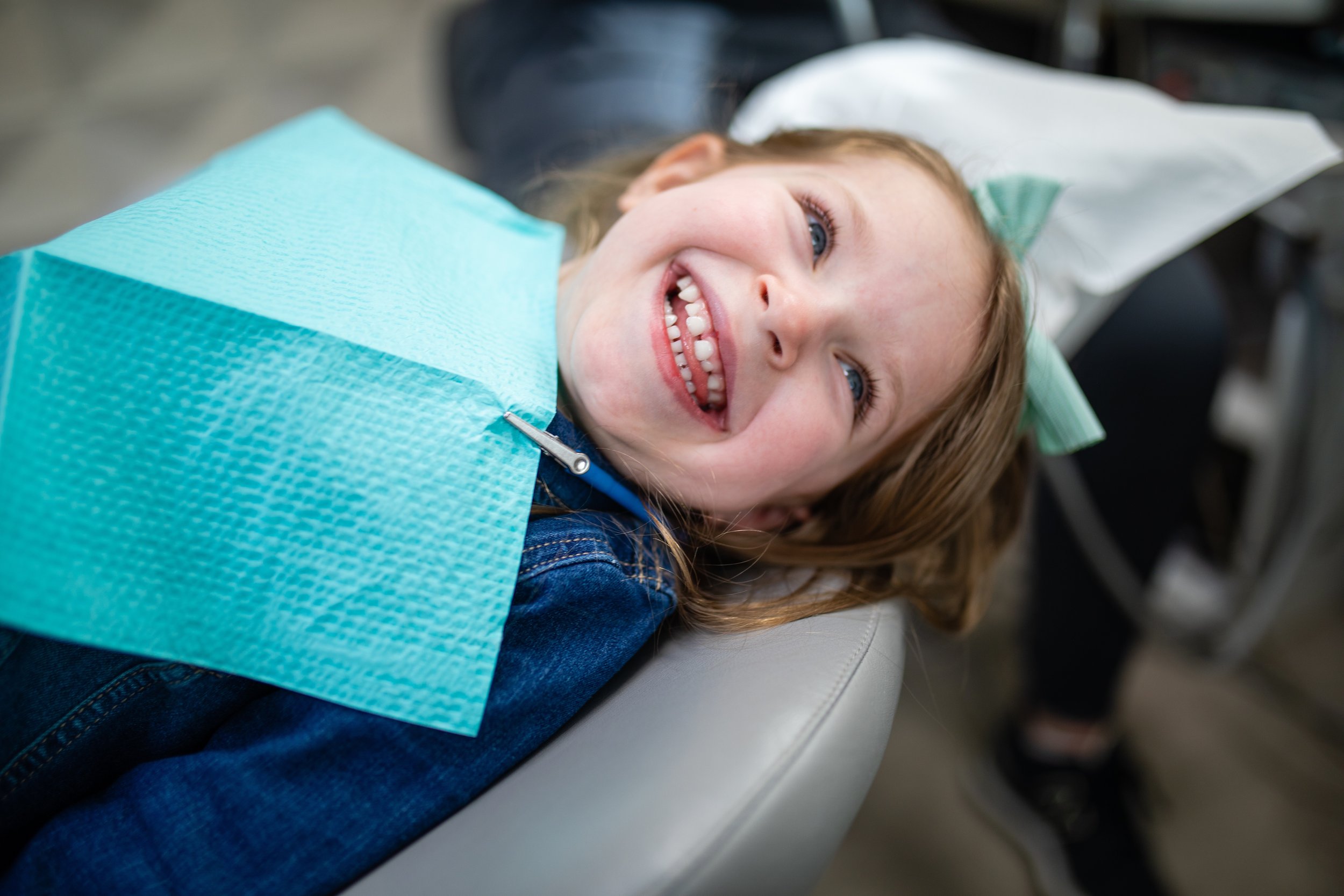 Young girl smiling happily at the dentist's chair, wearing a dental bib, with a green bow in her hair.