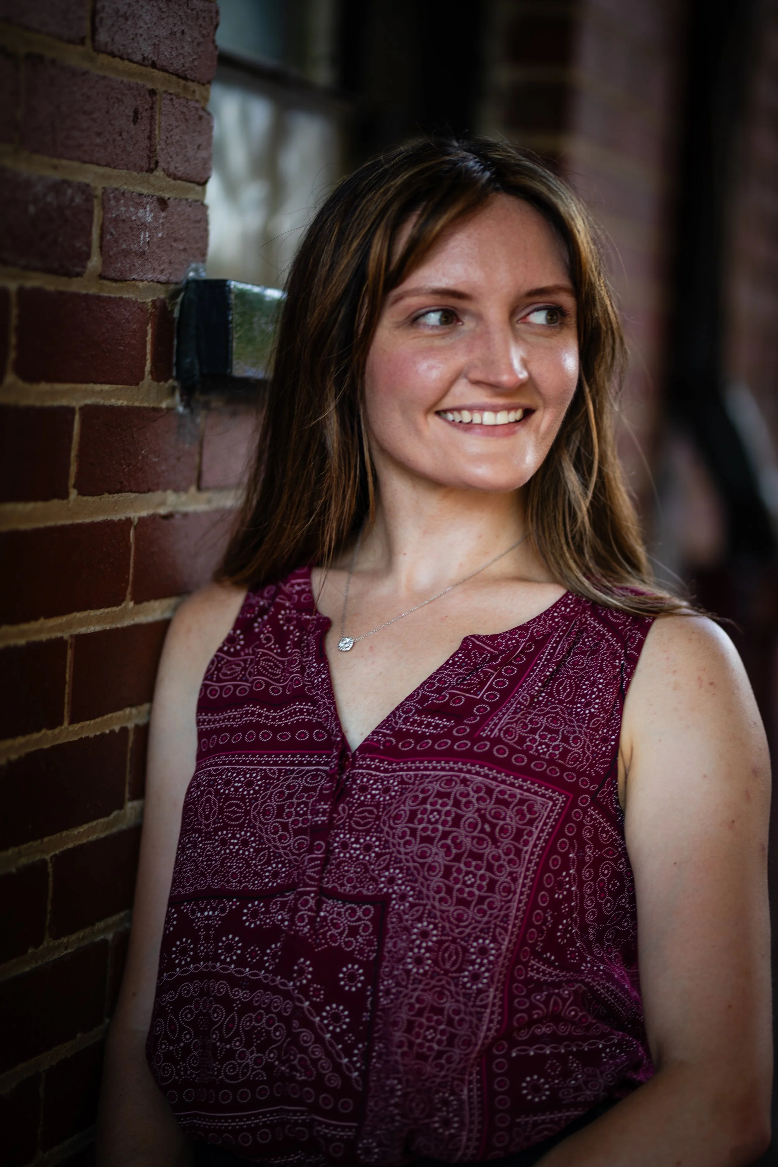 A woman with long brown hair smiling and looking to the side, standing against a brick wall with a window behind her.