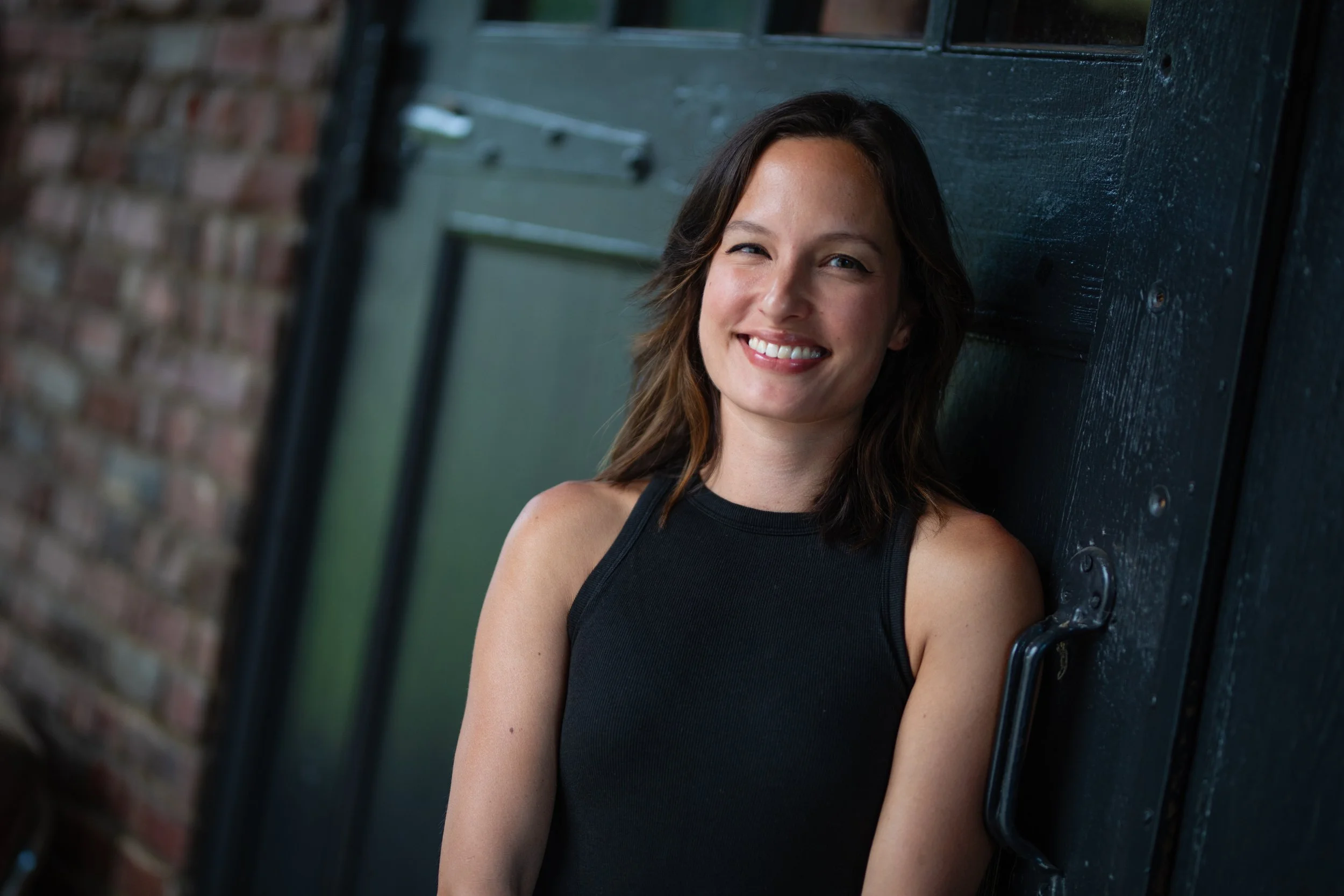 A woman with brown shoulder-length hair, wearing a black sleeveless top, smiling and leaning against a dark-colored door with a brick wall in the background.
