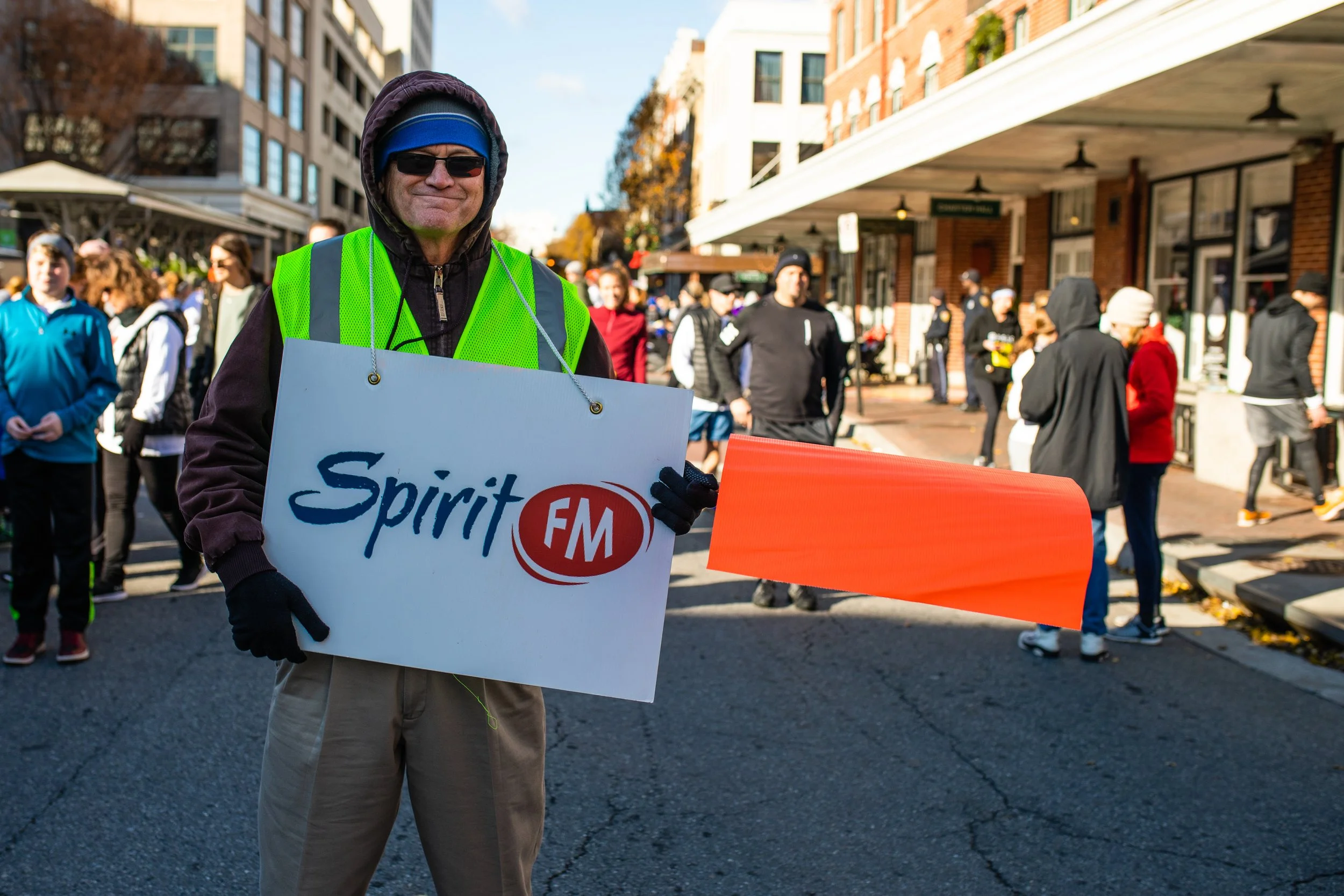 A man wearing sunglasses, a blue headband, a green safety vest, and gloves is holding a sign that reads "Spirit FM" during a street event or parade. There are several people in the background walking and standing on the sidewalk during daylight.