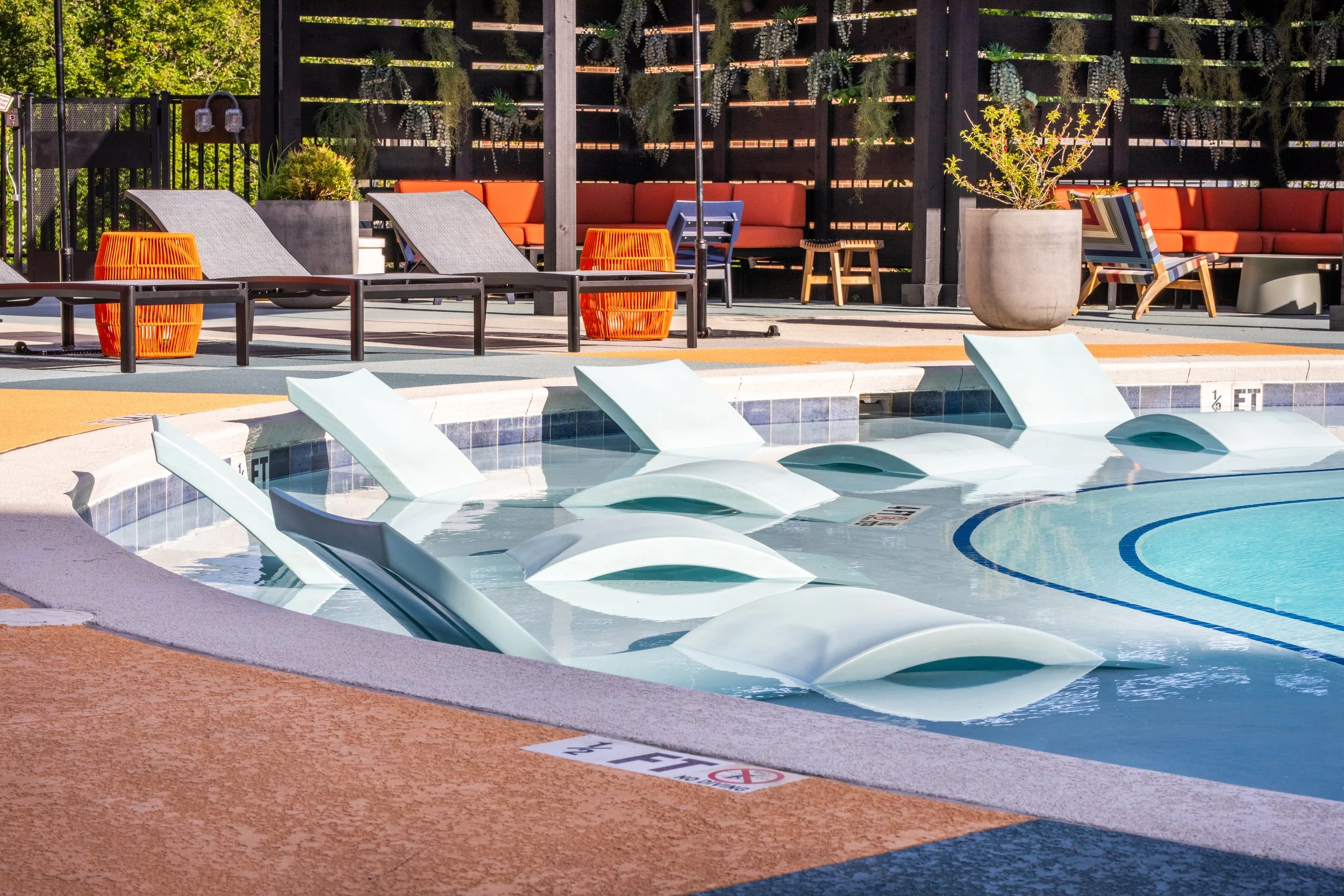 Empty swimming pool with multiple water slides, poolside lounge chairs, orange side tables, patio furniture, potted plants, and a wooden fence with hanging plants in the background.
