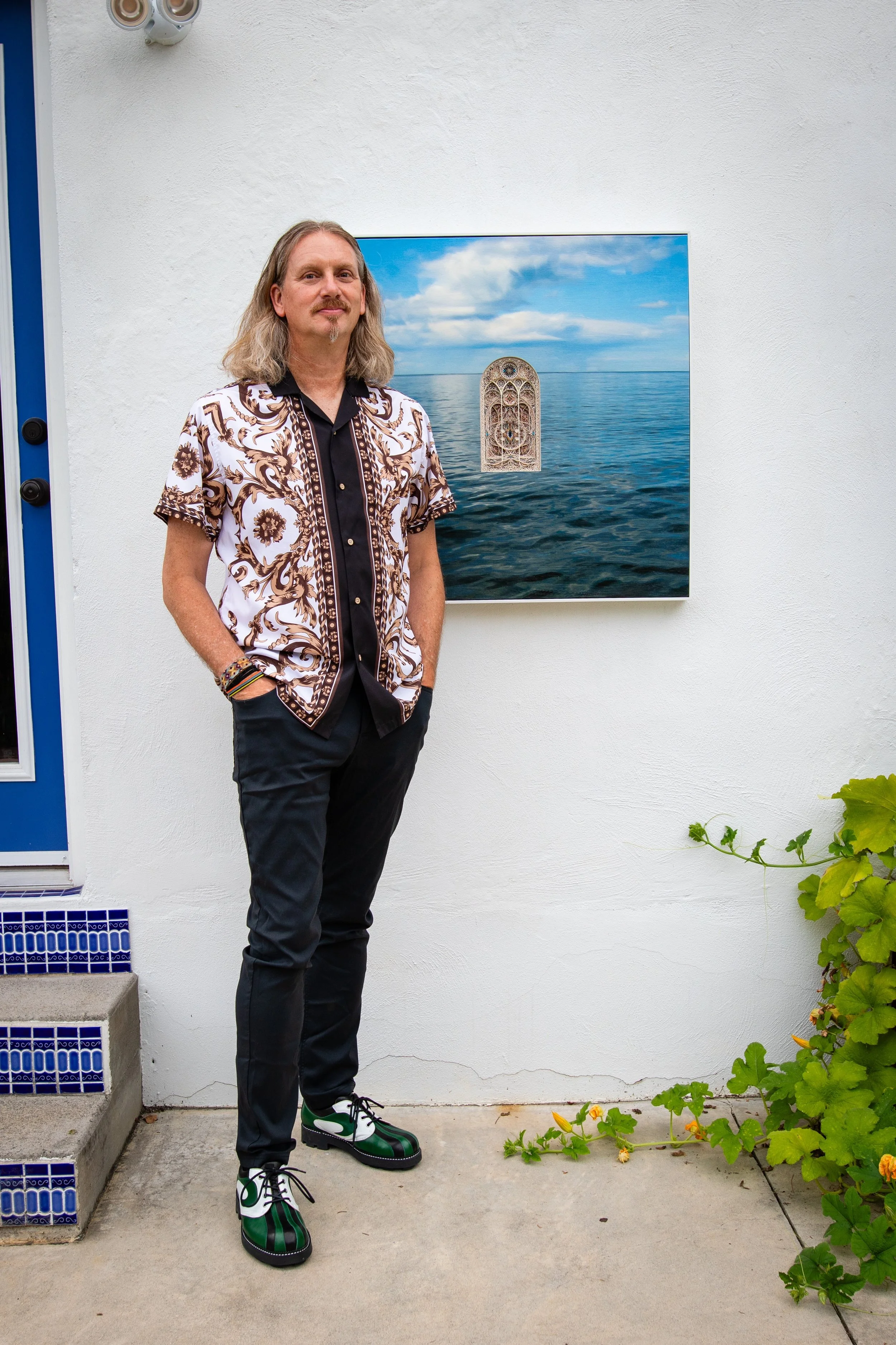 A man with long hair and a goatee standing outside near a painting of the ocean on the wall behind him. The man is wearing a patterned short-sleeve shirt, black pants, and colorful shoes. There are green plants on the ground next to him and blue tile