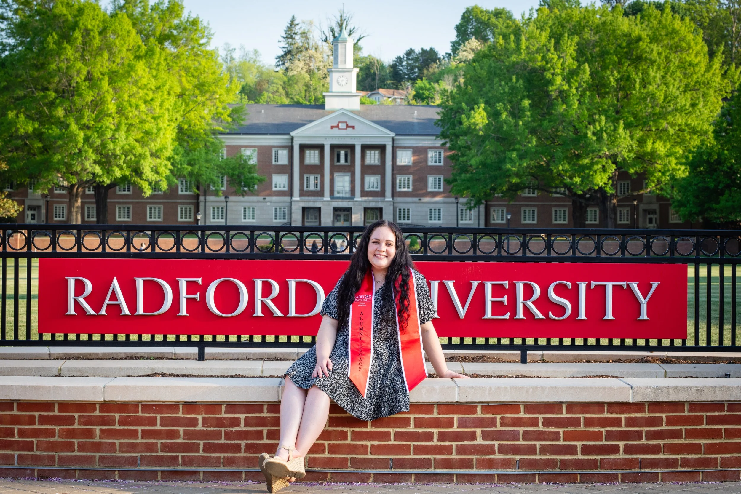 A young woman with long dark hair sitting on a brick ledge in front of a red sign that reads 'Radford University.' She is smiling, wearing a black and white dress, and has orange graduation sashes around her neck. The background includes green trees 