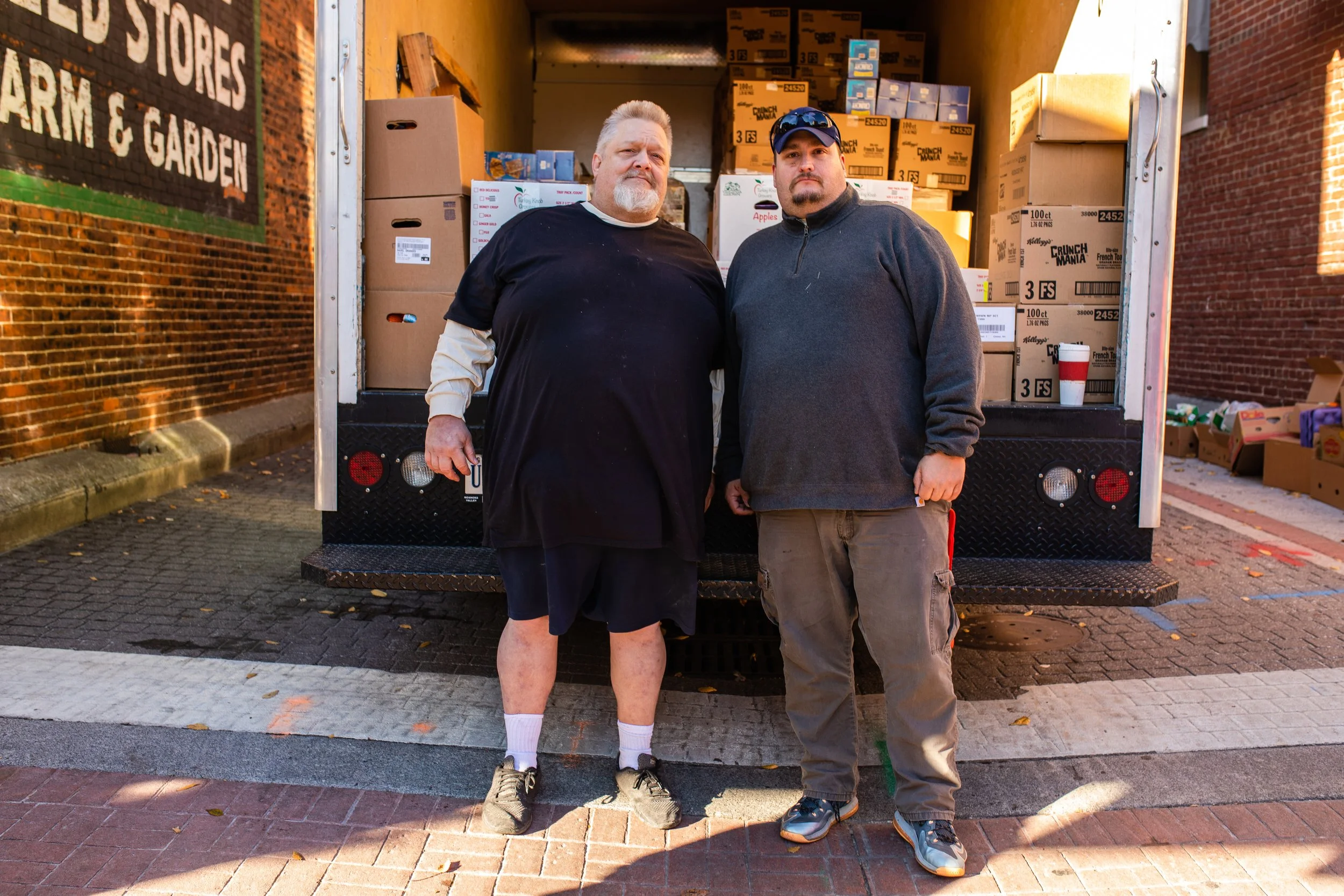 Two men standing in front of a moving truck loaded with boxes, outdoors on a sidewalk during daytime.