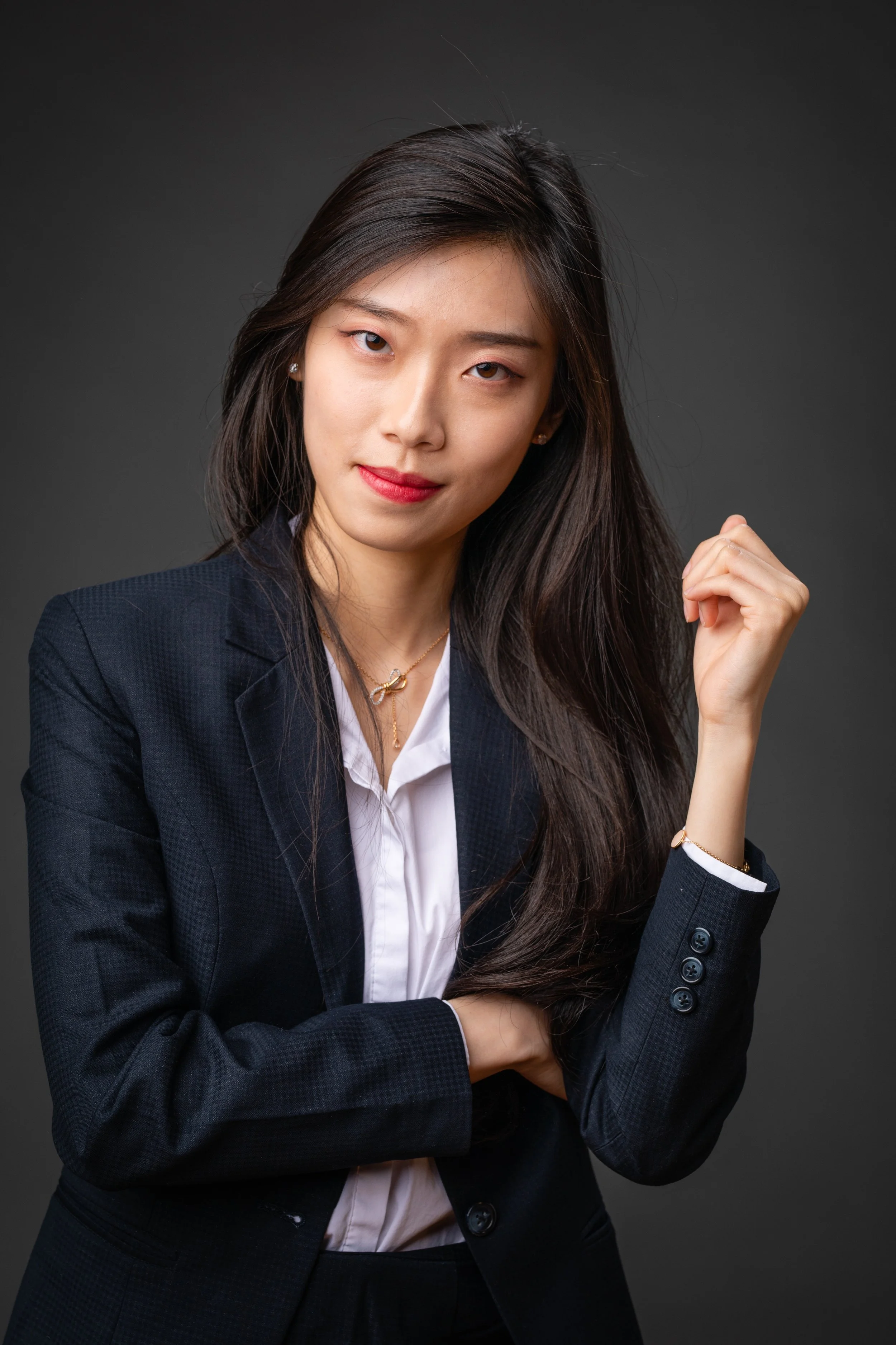 A confident young woman with long dark hair, wearing a black blazer and white shirt, posing against a dark background.