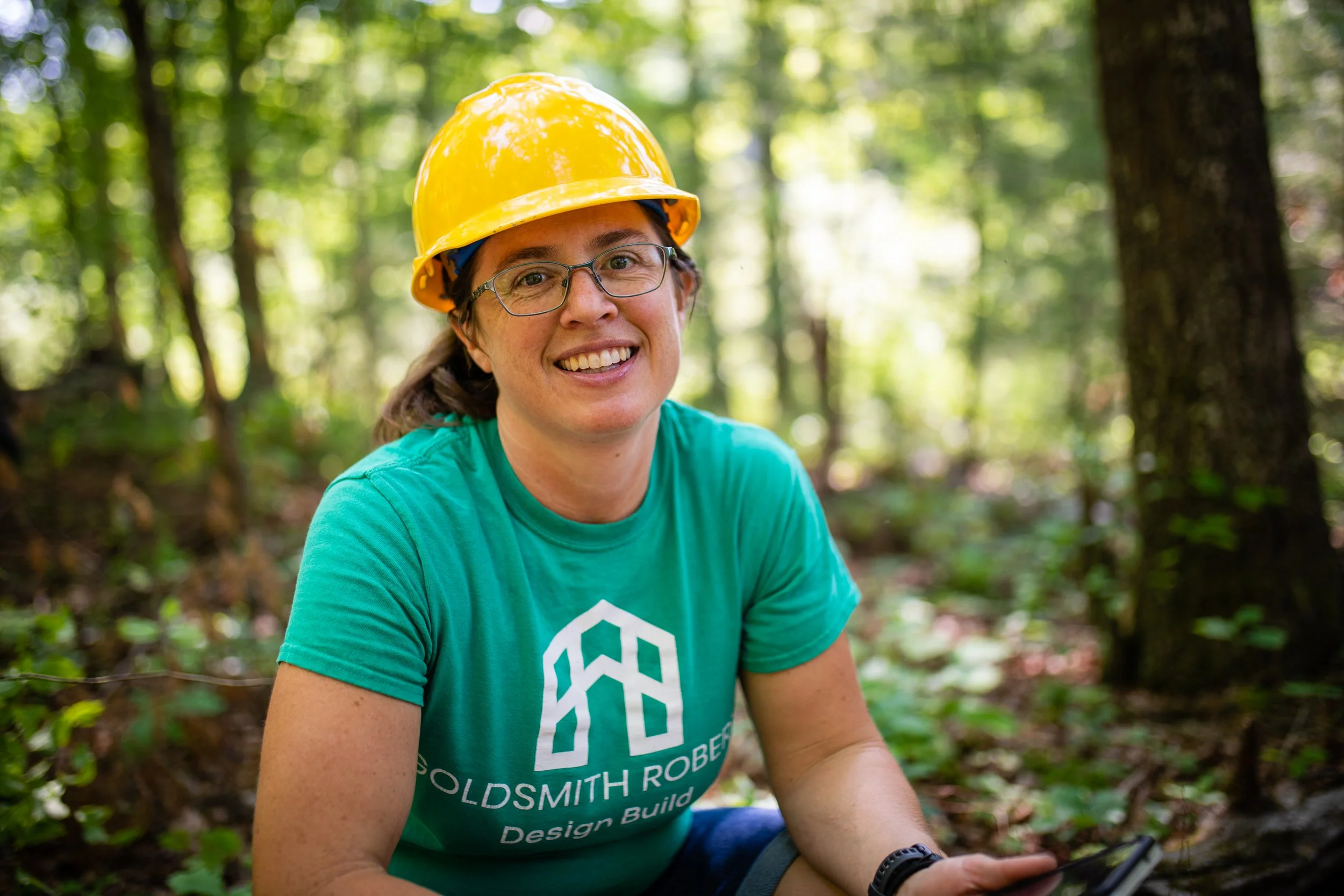 A woman wearing glasses, a yellow hard hat, and a teal T-shirt with the logo 'Oldsmth Robe Design Build' is smiling in a forested area during daytime.