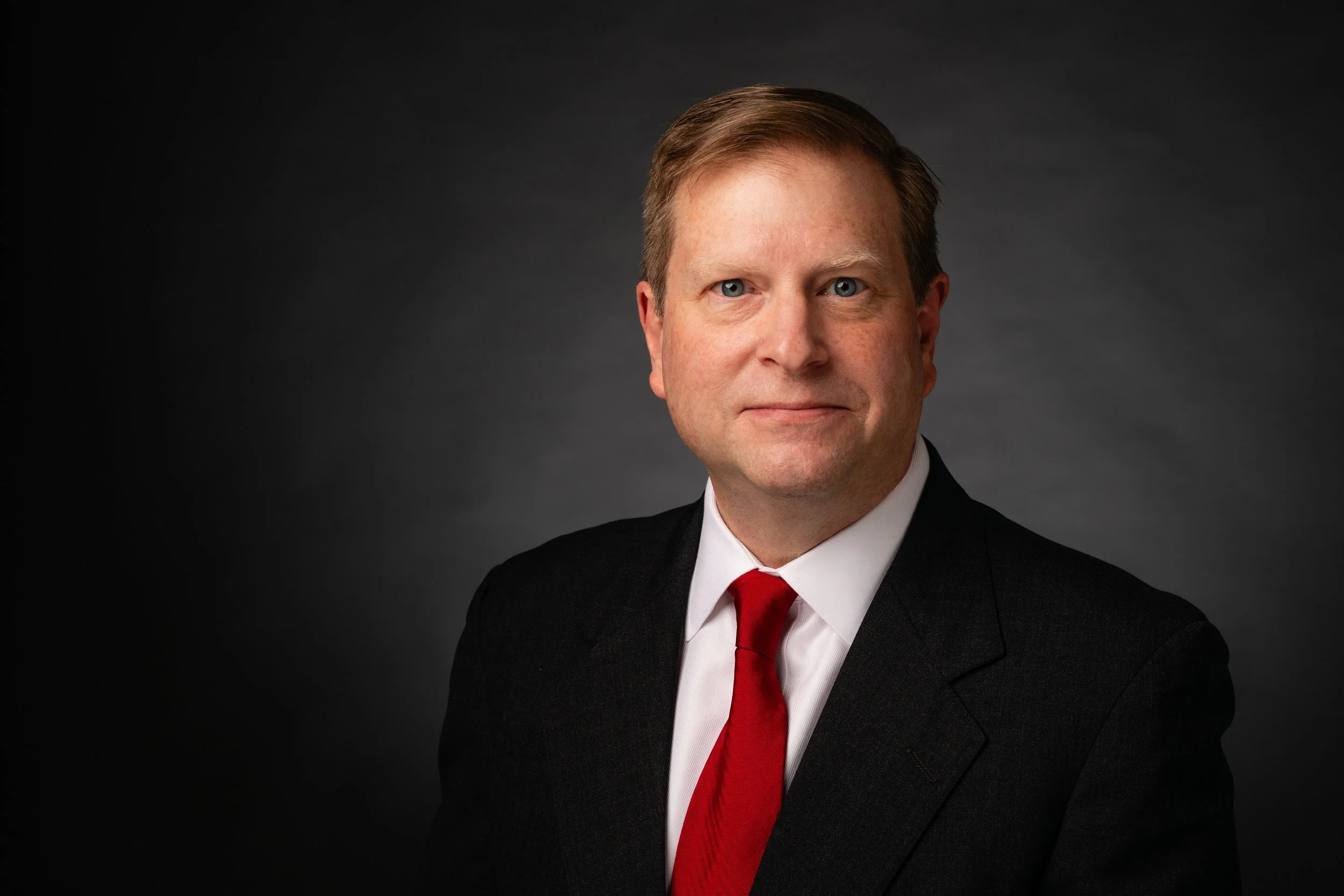 Professional portrait of a man wearing a suit, white shirt, and red tie against a dark background.