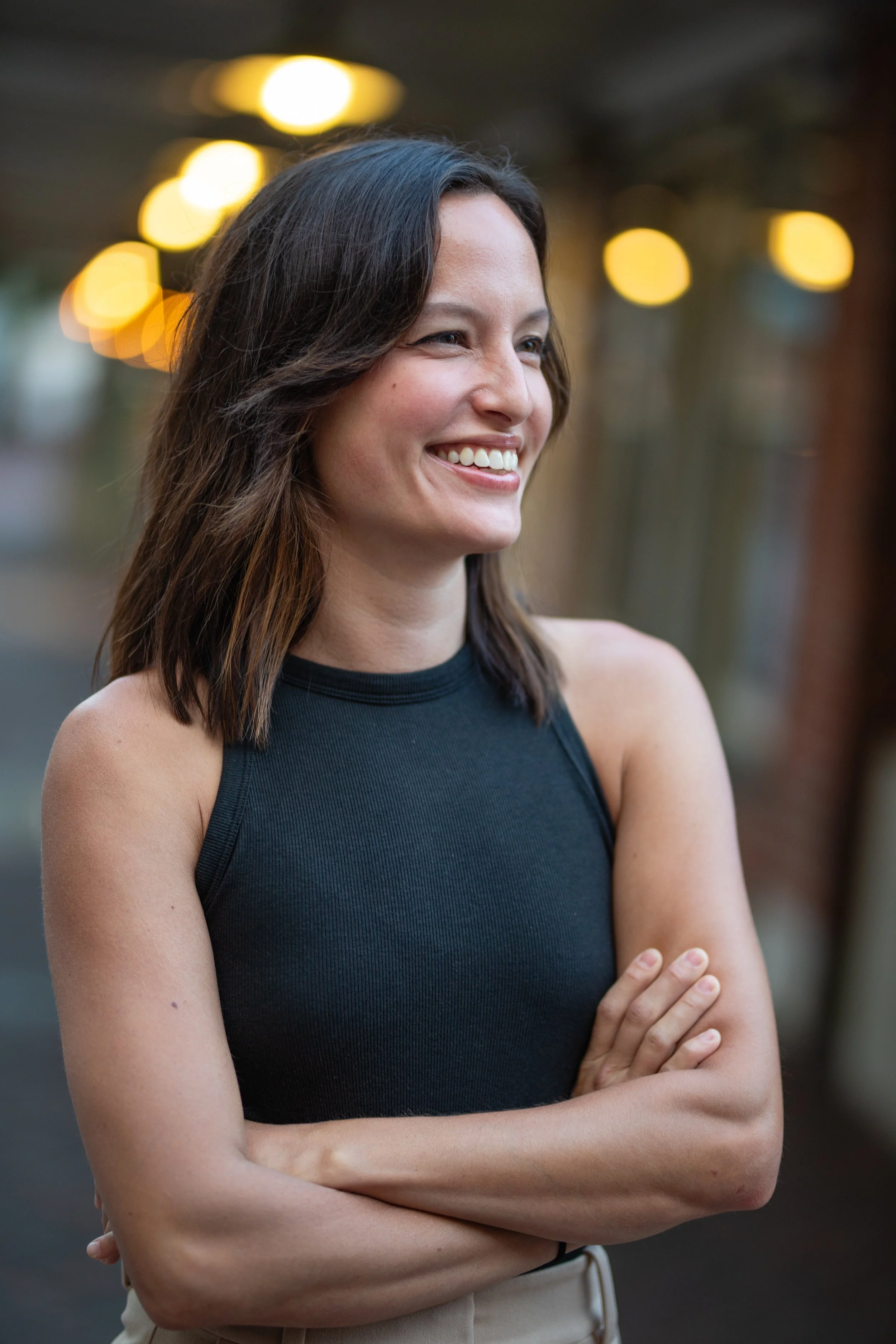 A woman with shoulder-length brown hair smiling outdoors at dusk, wearing a sleeveless black top with arms crossed.