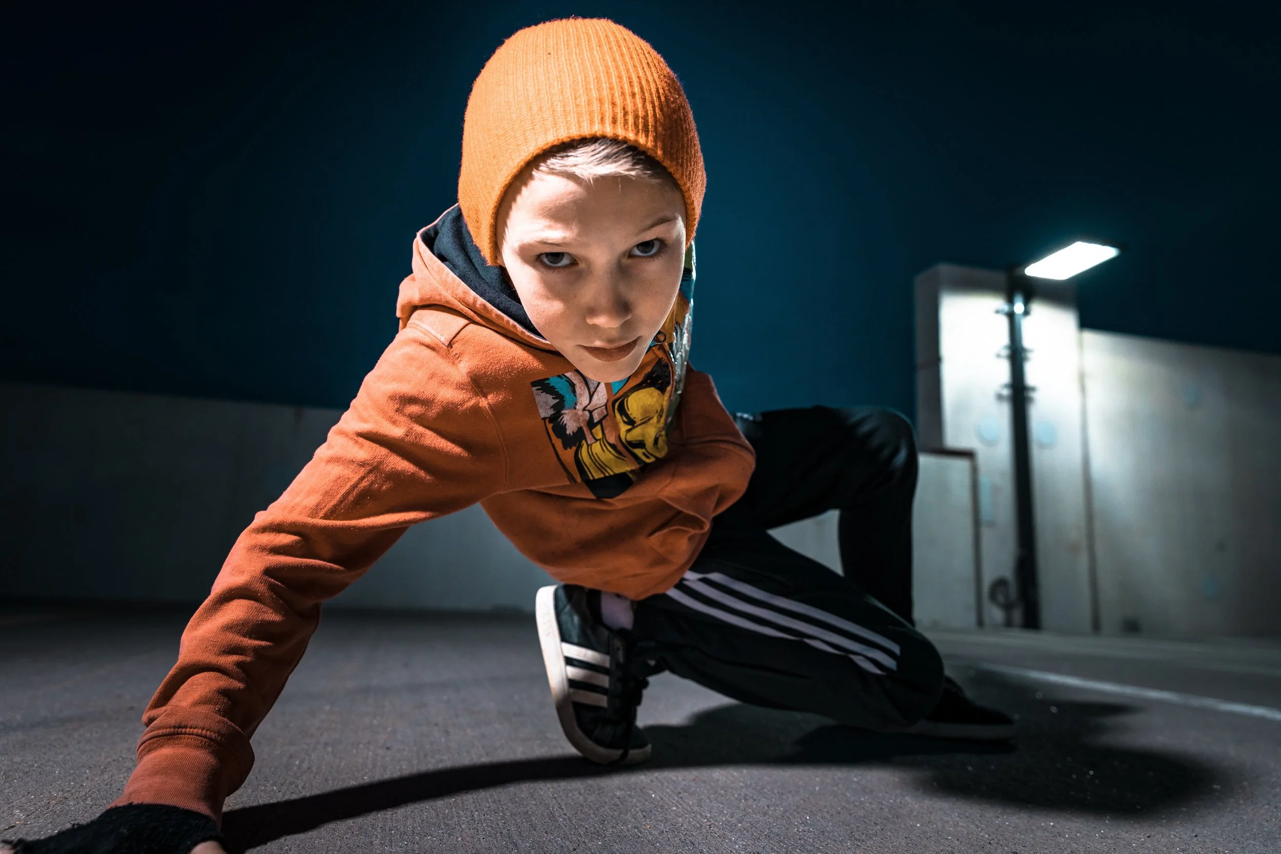 A young boy in an orange beanie and jacket crouches on the ground in a dark parking garage, looking intensely at the camera.
