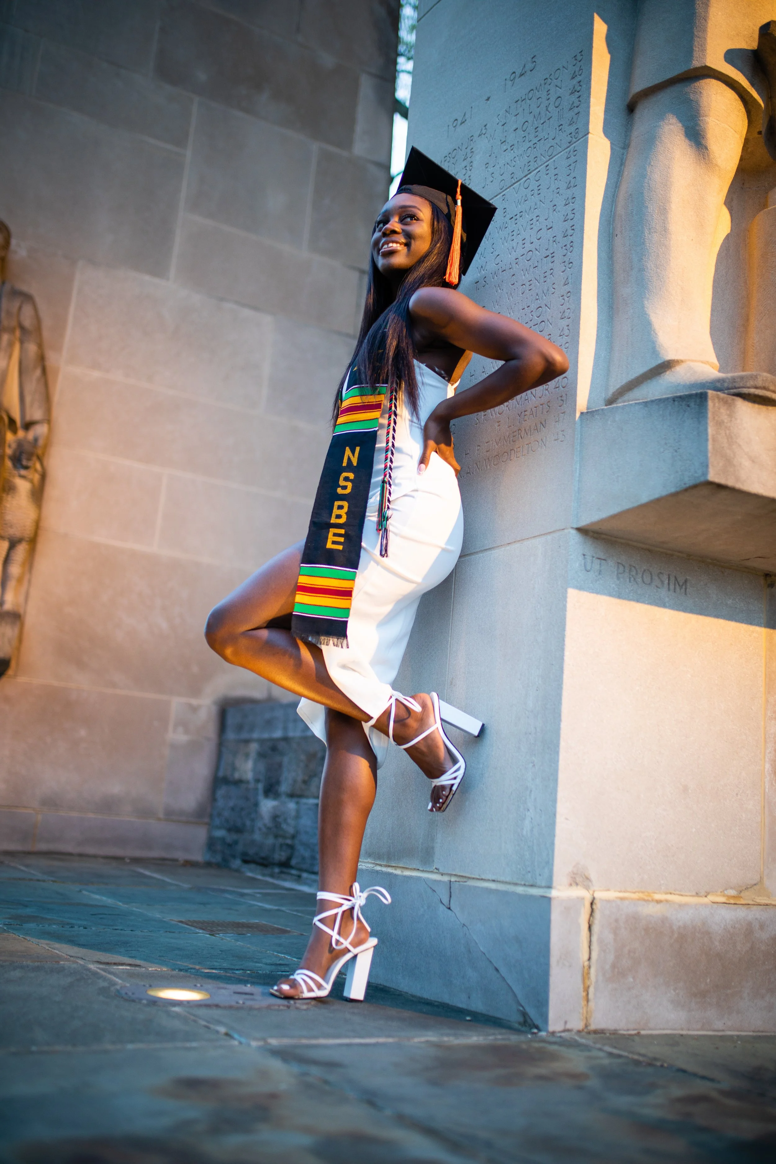 A happy young woman in a white dress and white high-heeled sandals is posing outdoors, leaning against a stone monument with inscriptions. She is wearing a graduation cap and stole, celebrating her graduation.