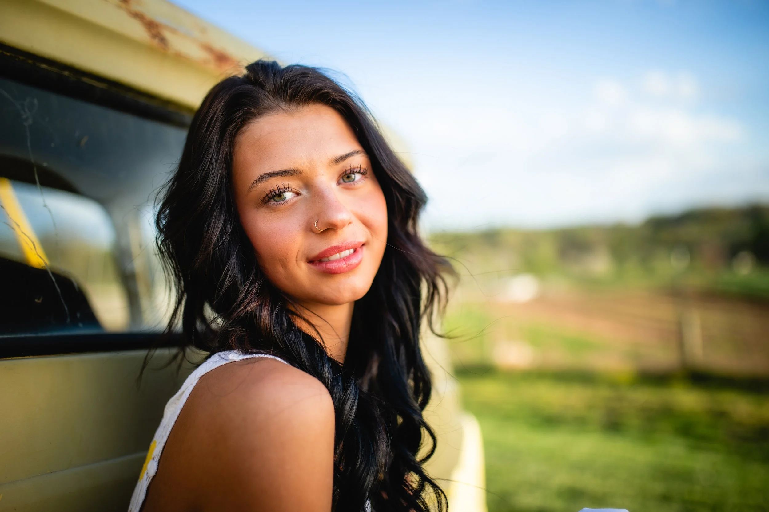A young woman with long dark hair and a nose ring smiles while leaning against a yellow vehicle outdoors, with a blurred green landscape and blue sky in the background.