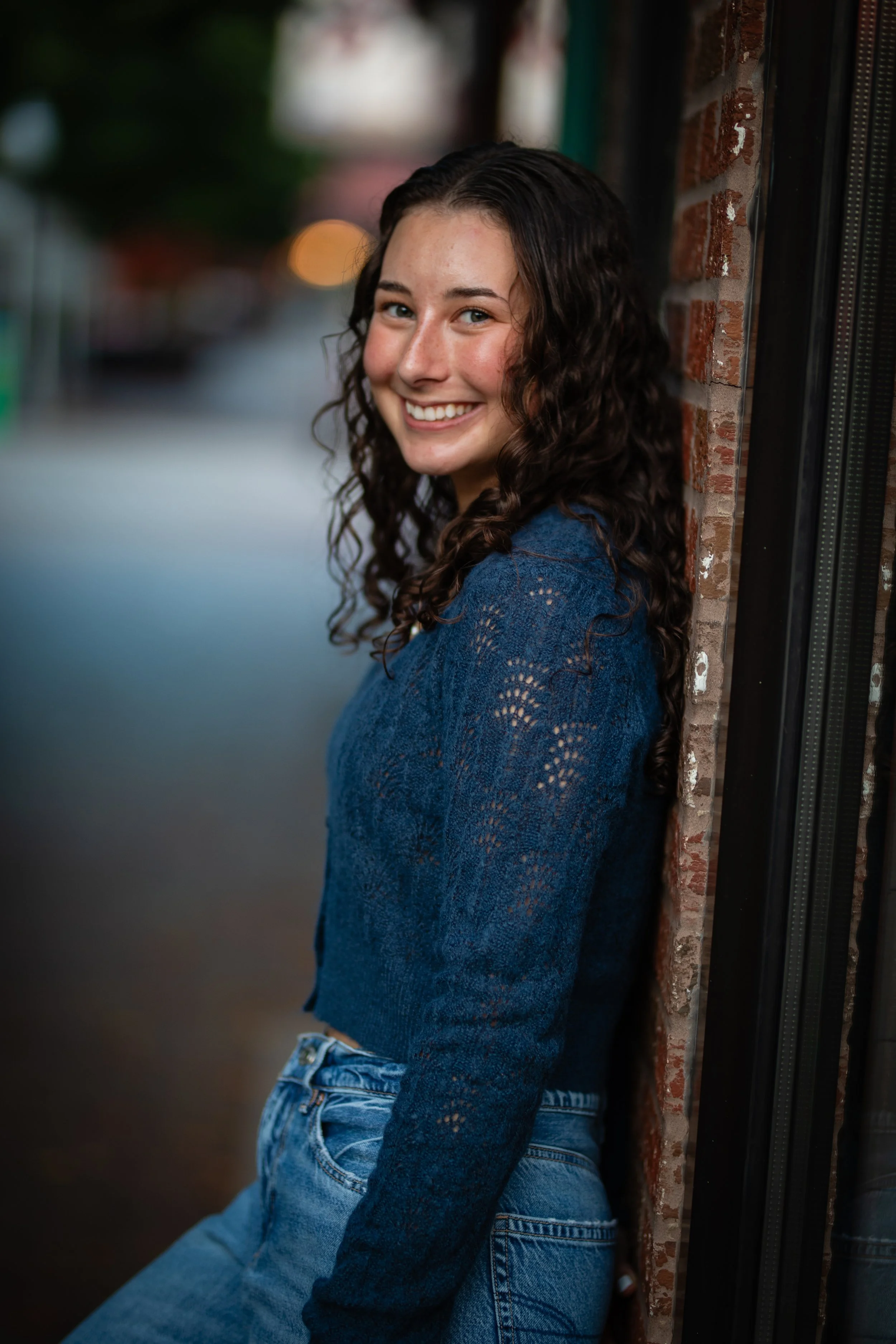 A young woman with curly dark hair smiling and leaning against a brick wall outdoors.