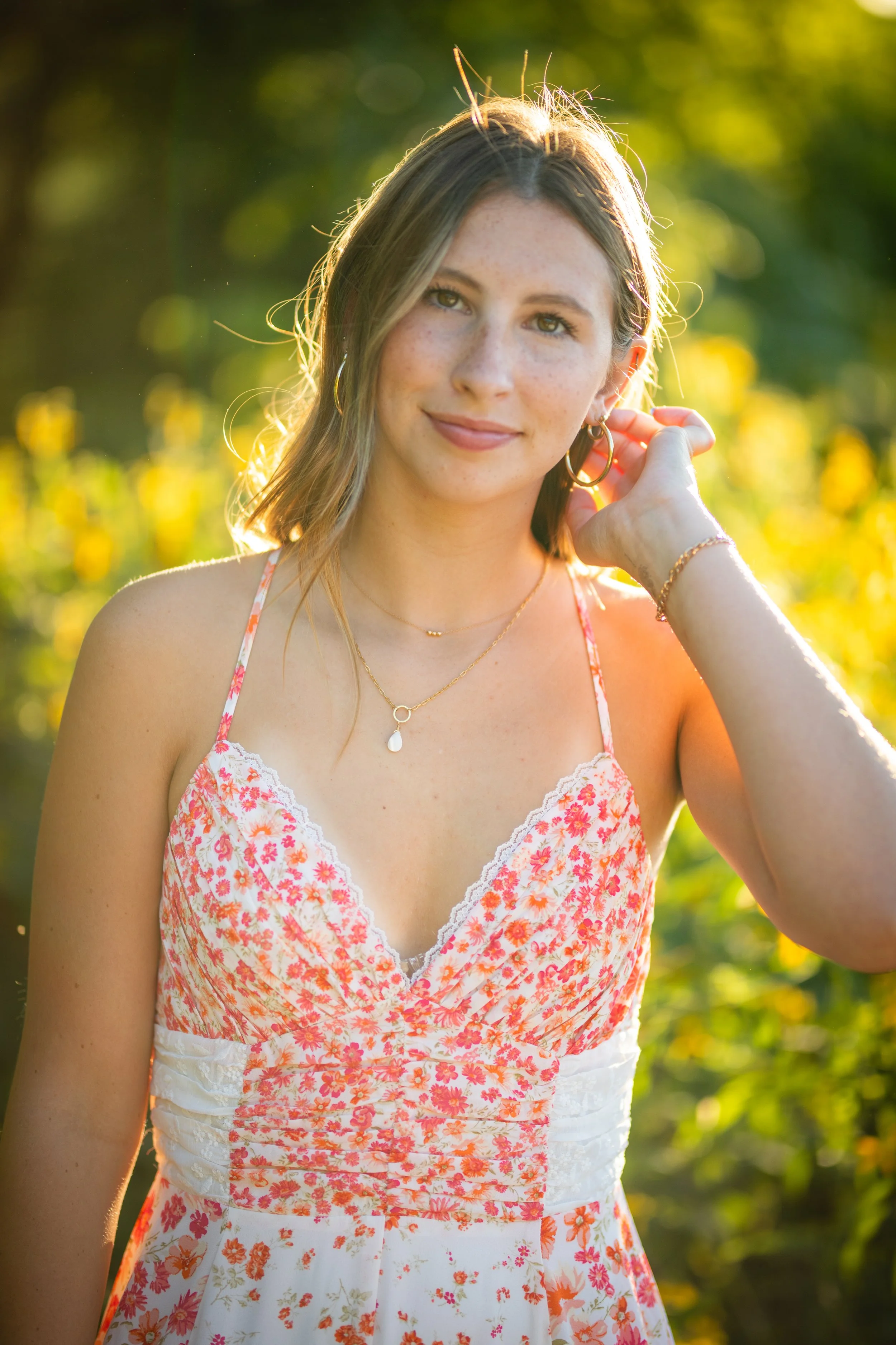 A young woman with light brown hair and freckles standing outdoors in a field of yellow flowers during golden hour. She is wearing a floral dress with thin straps, gold jewelry, and has a gentle smile, with sunlight illuminating her face.