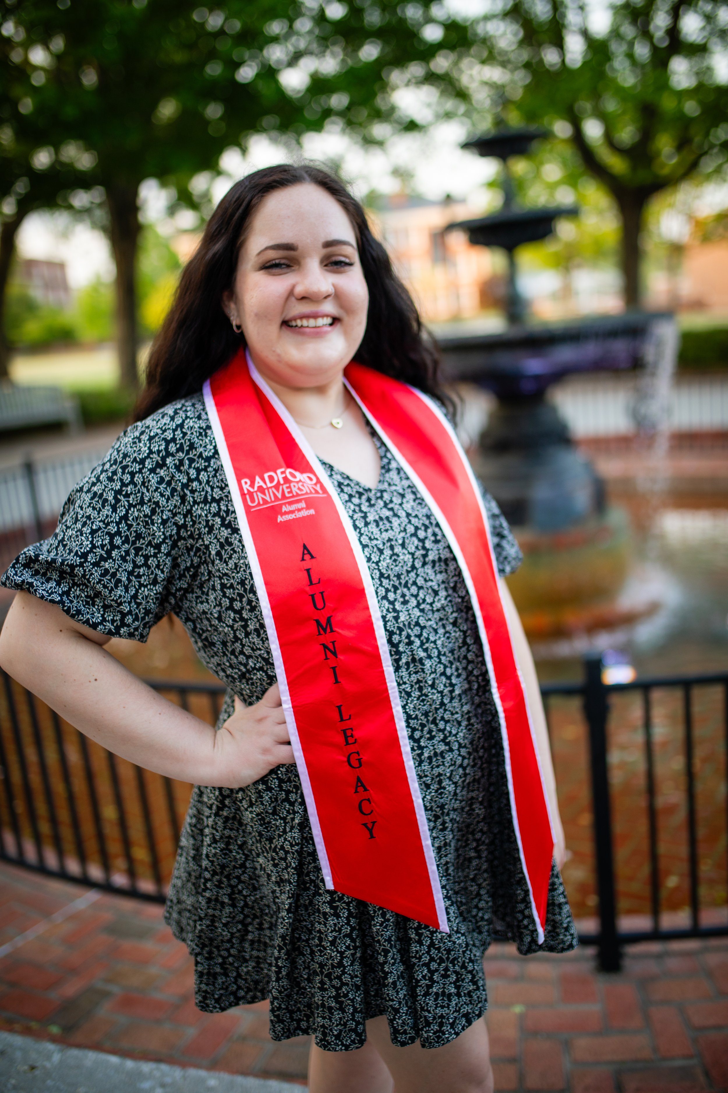 A young woman smiling outdoors wearing a red sash that says 'Alumni Legacy' and a black and white patterned dress, with trees and a fountain in the background.