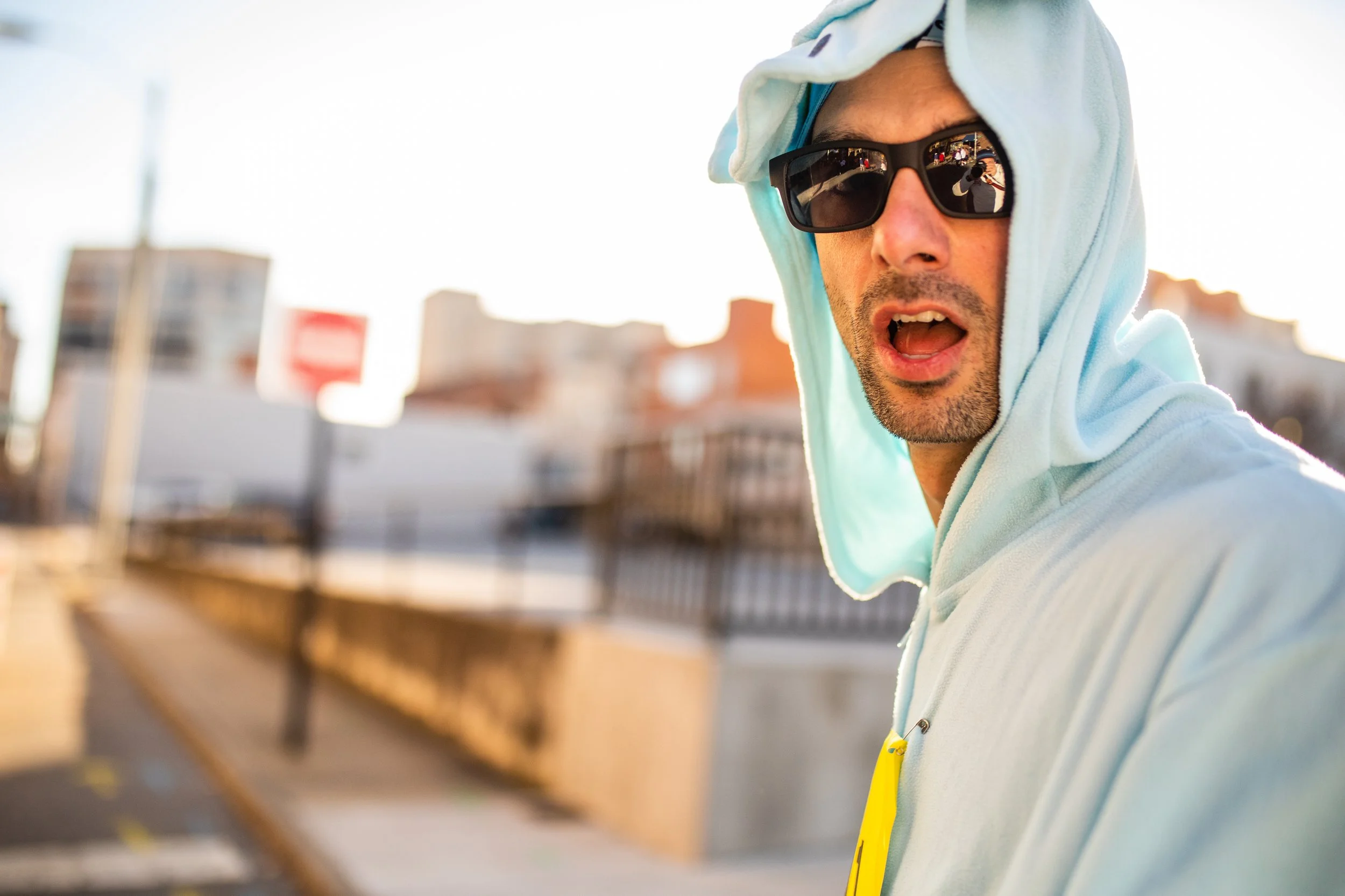 A man wearing sunglasses and a light blue hoodie with the hood up, standing outdoors with a blurred cityscape background.