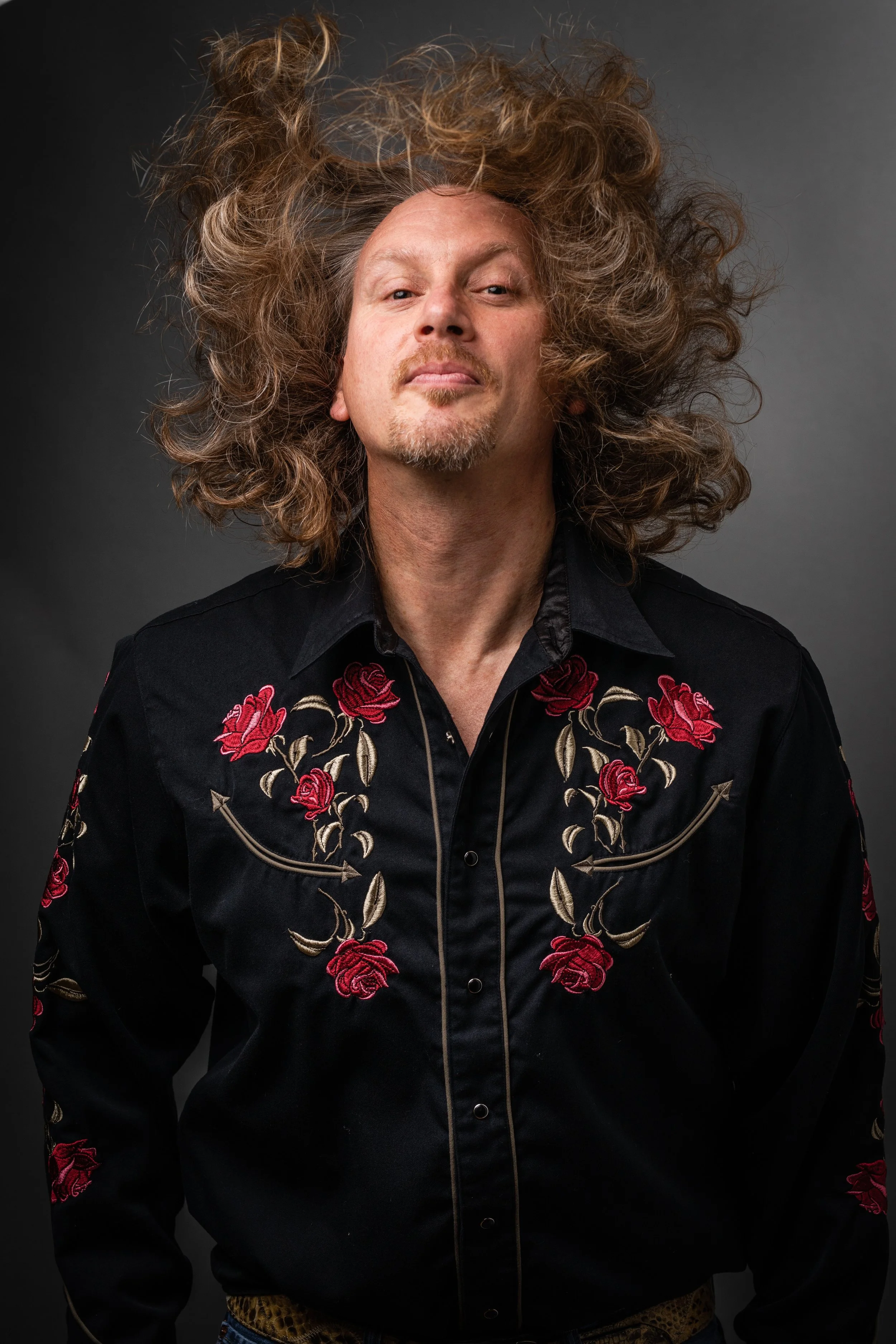 A man with curly, voluminous hair wearing a black western-style shirt with embroidered red roses and leaves, against a dark gray background.