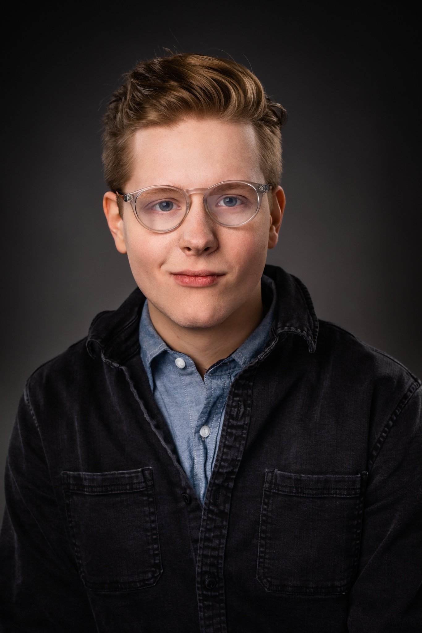 Portrait of a young man with glasses and styled hair, wearing a blue shirt and black jacket, against a dark background.