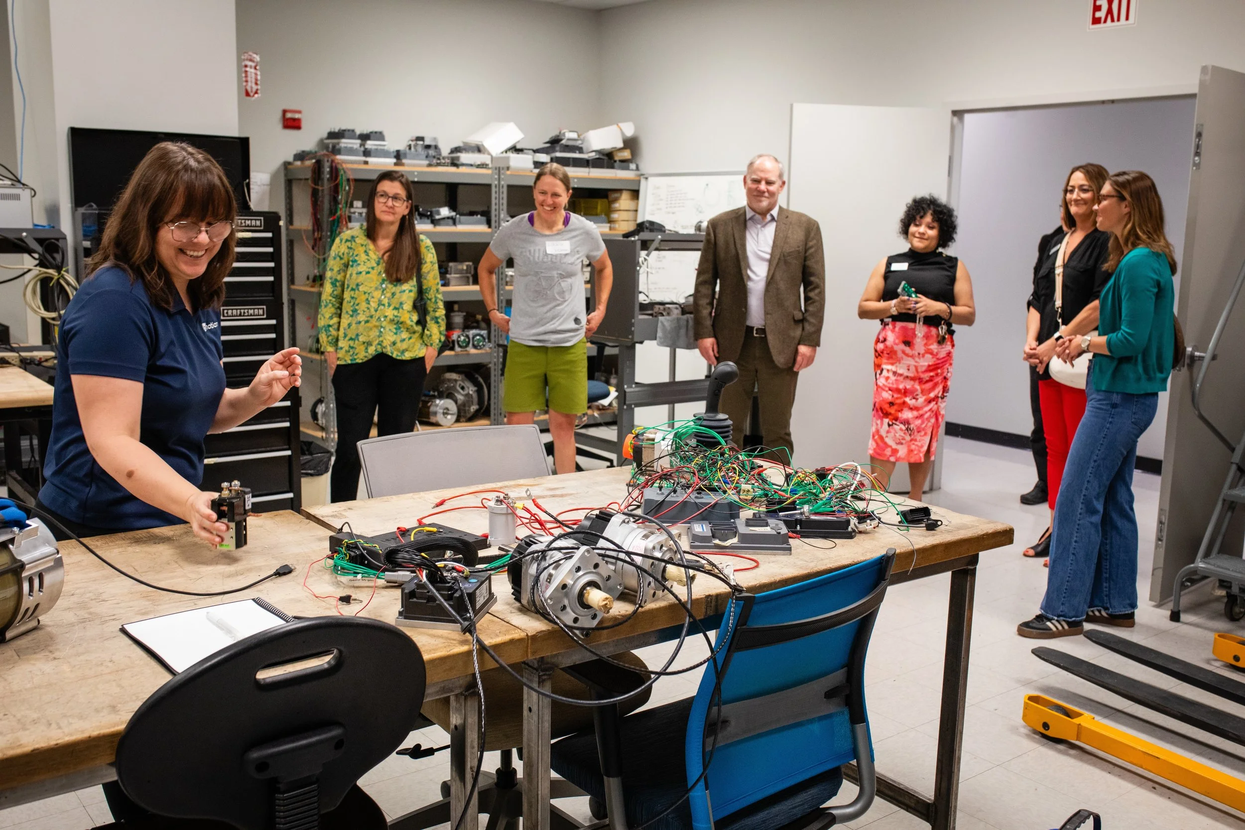 Group of people in a laboratory around a table with electrical and mechanical components, some smiling and observing a woman demonstrating a project.
