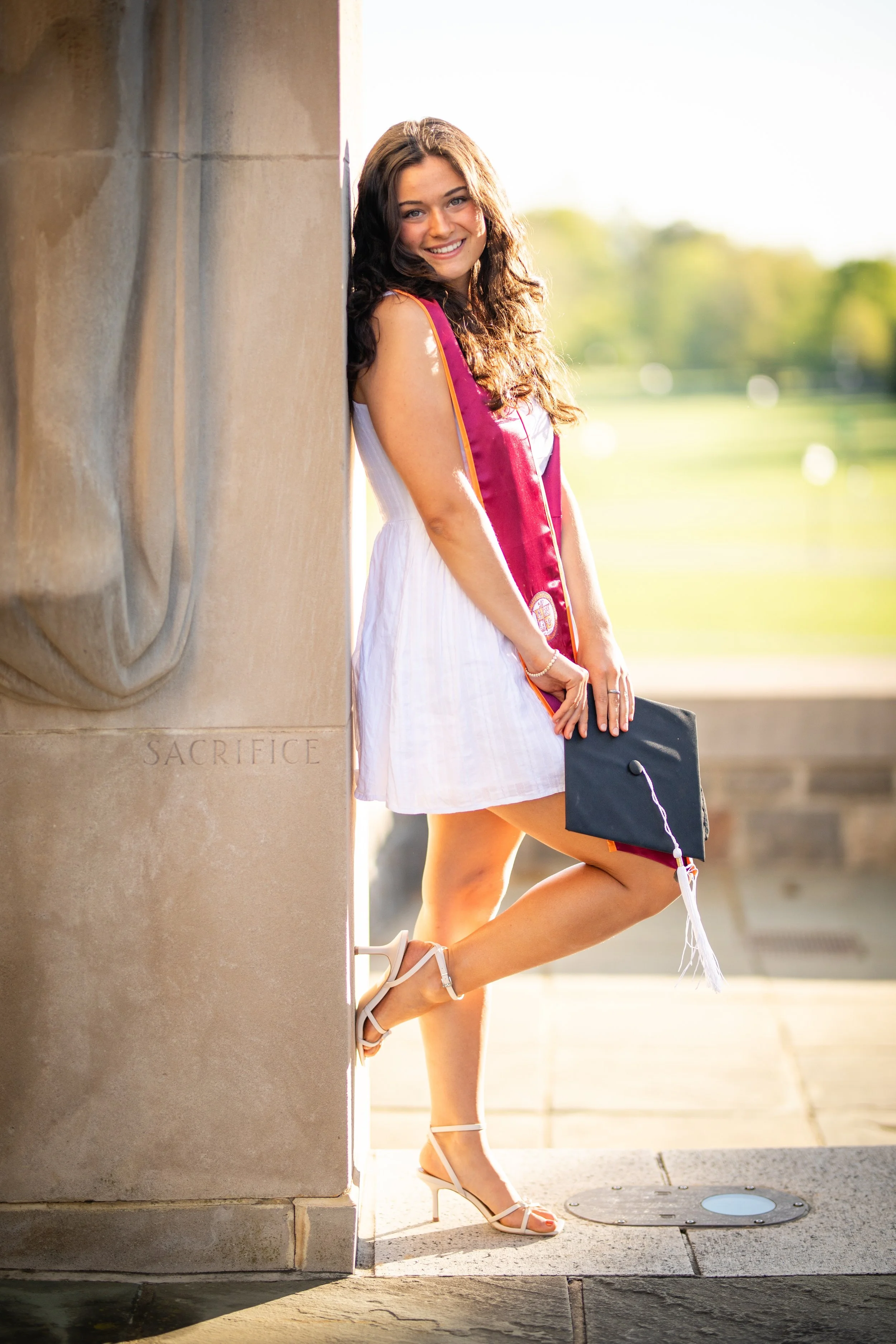 Young woman in a white dress wearing a graduation stole and cap, standing outdoors and smiling.