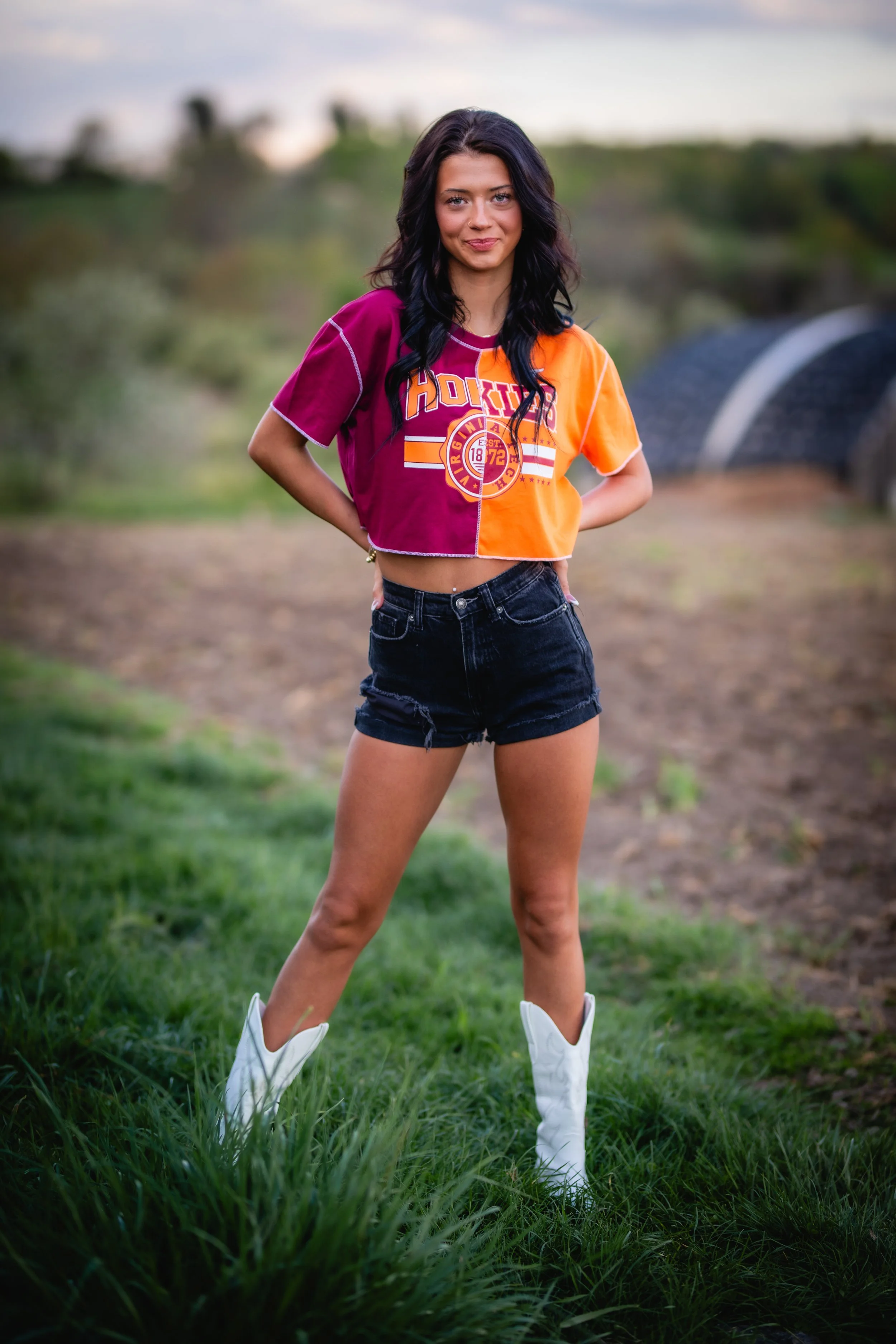 Young woman standing outdoors on grass, wearing a colorful crop top, black shorts, and white cowboy boots, with a blurred rural background.