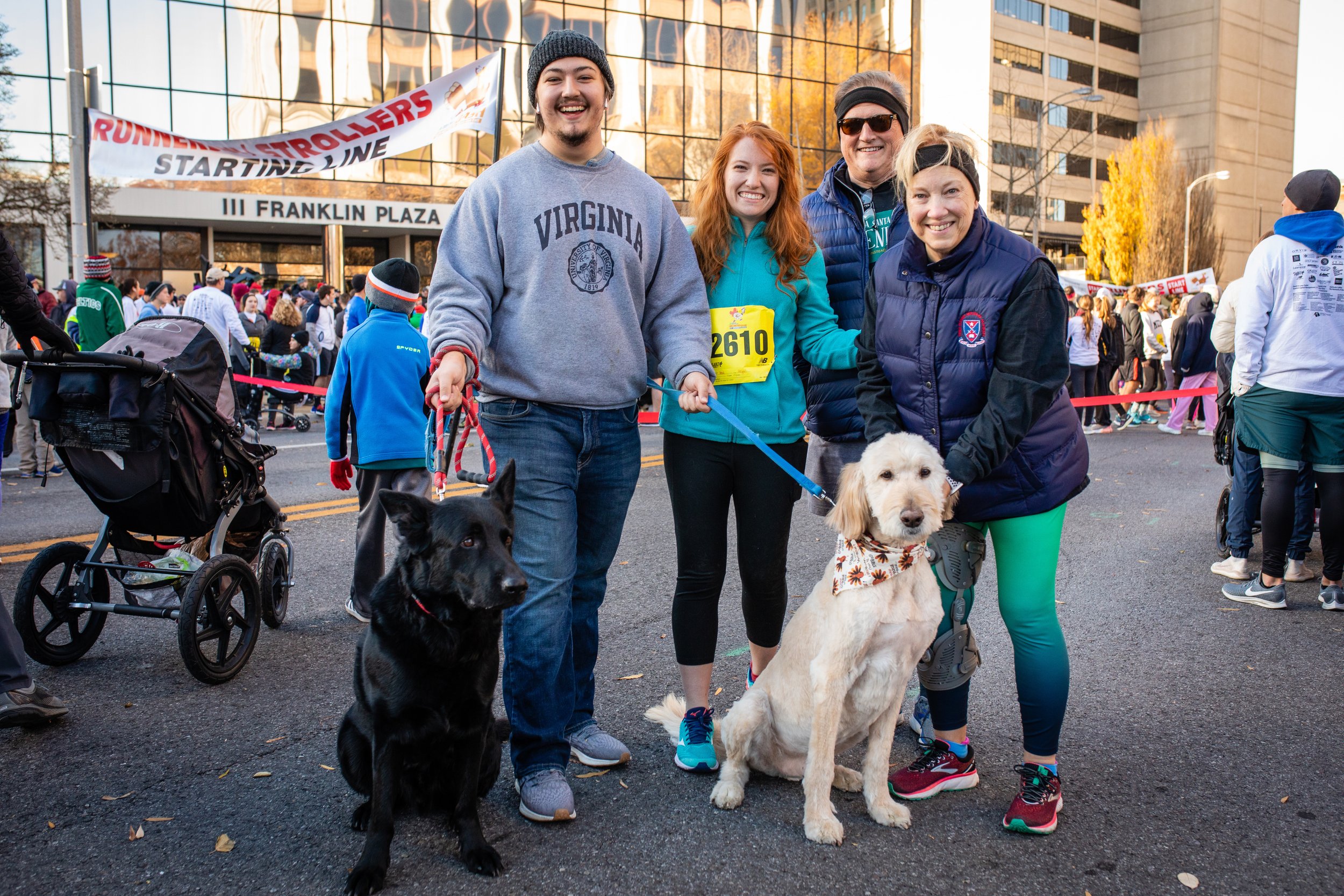 Group of four smiling people standing together at an outdoor event, with two dogs, at the start line of a race in a city setting.