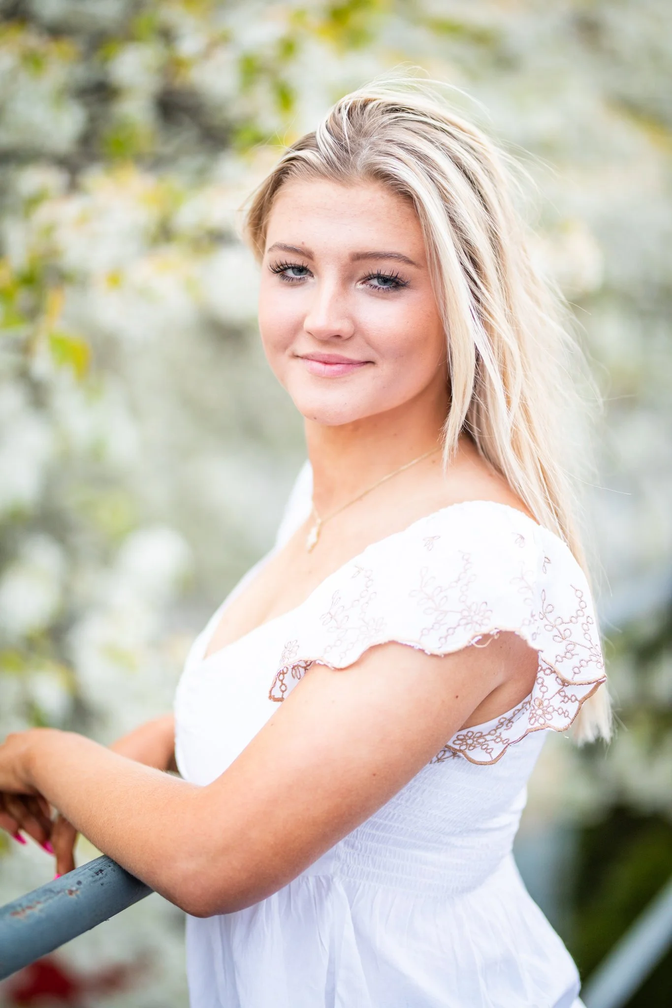 Portrait of a young woman with blonde hair wearing a white dress with lace detailing on the sleeves, standing outdoors near greenery.