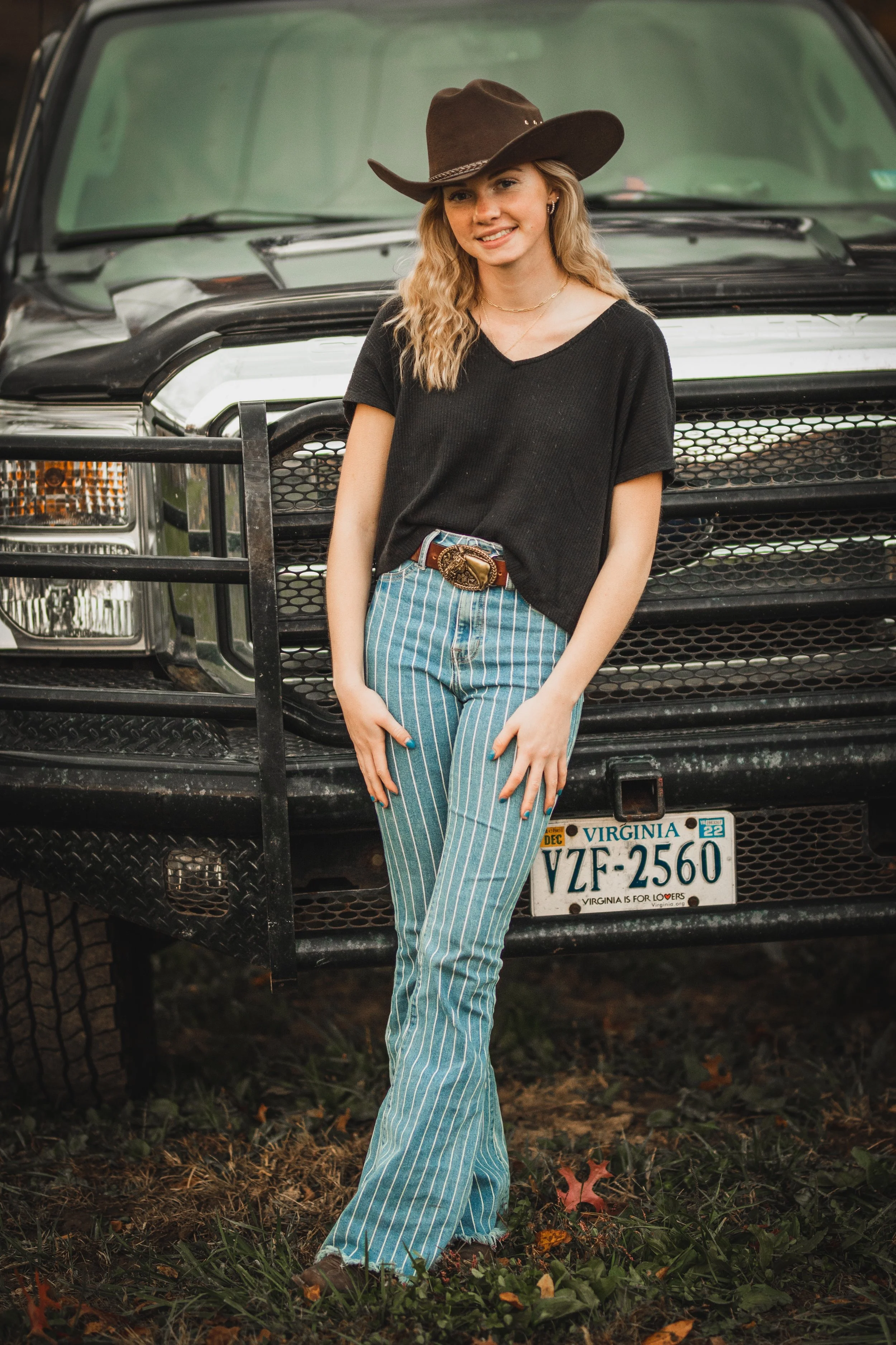 A young woman standing in front of a black truck with Virginia license plates, wearing a black t-shirt, striped blue and white bell-bottom jeans, a brown cowboy hat, and a brown belt, smiling at the camera.