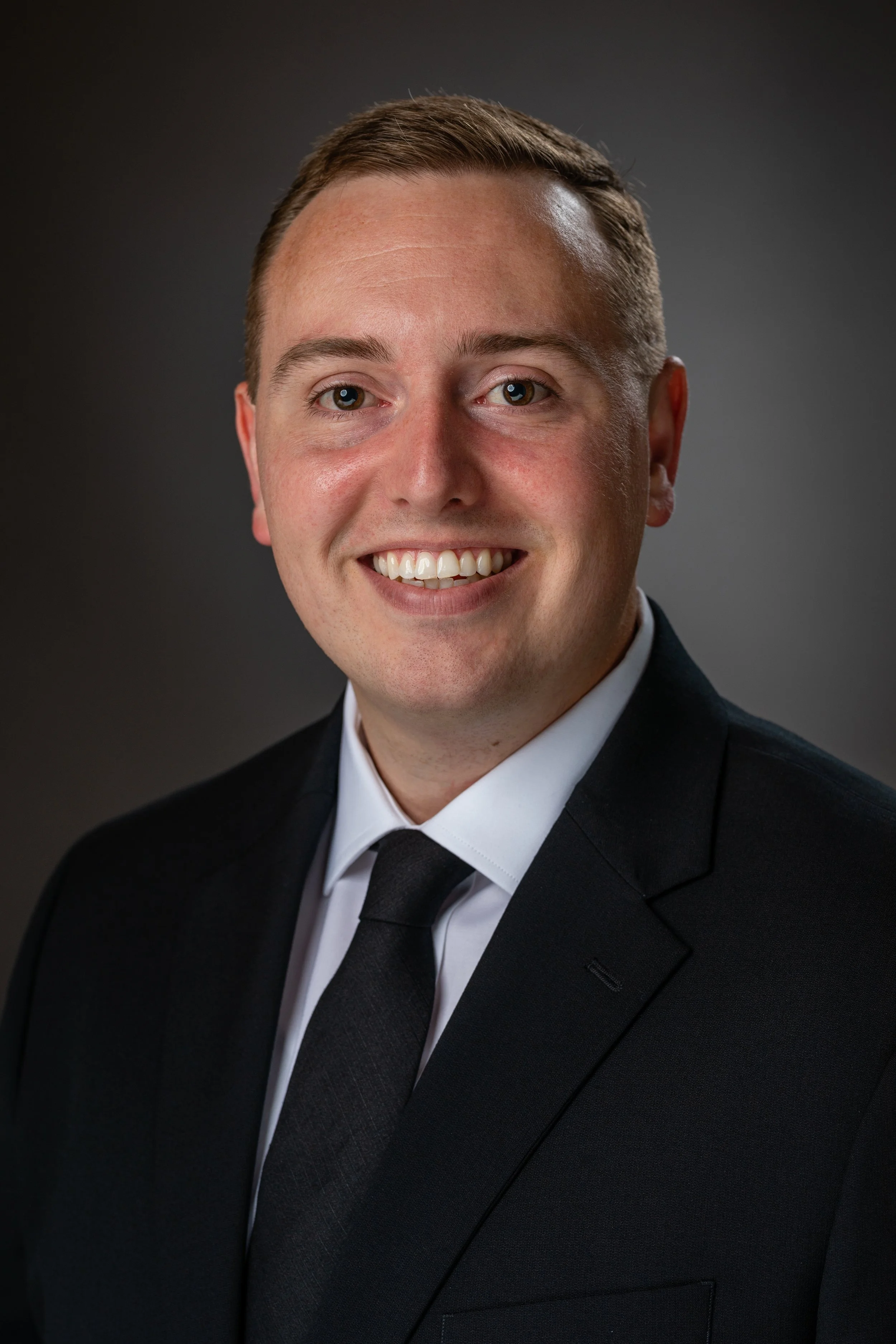 A professional headshot of a young man in a black suit, white shirt, and black tie, smiling with short light brown hair against a dark gradient background.